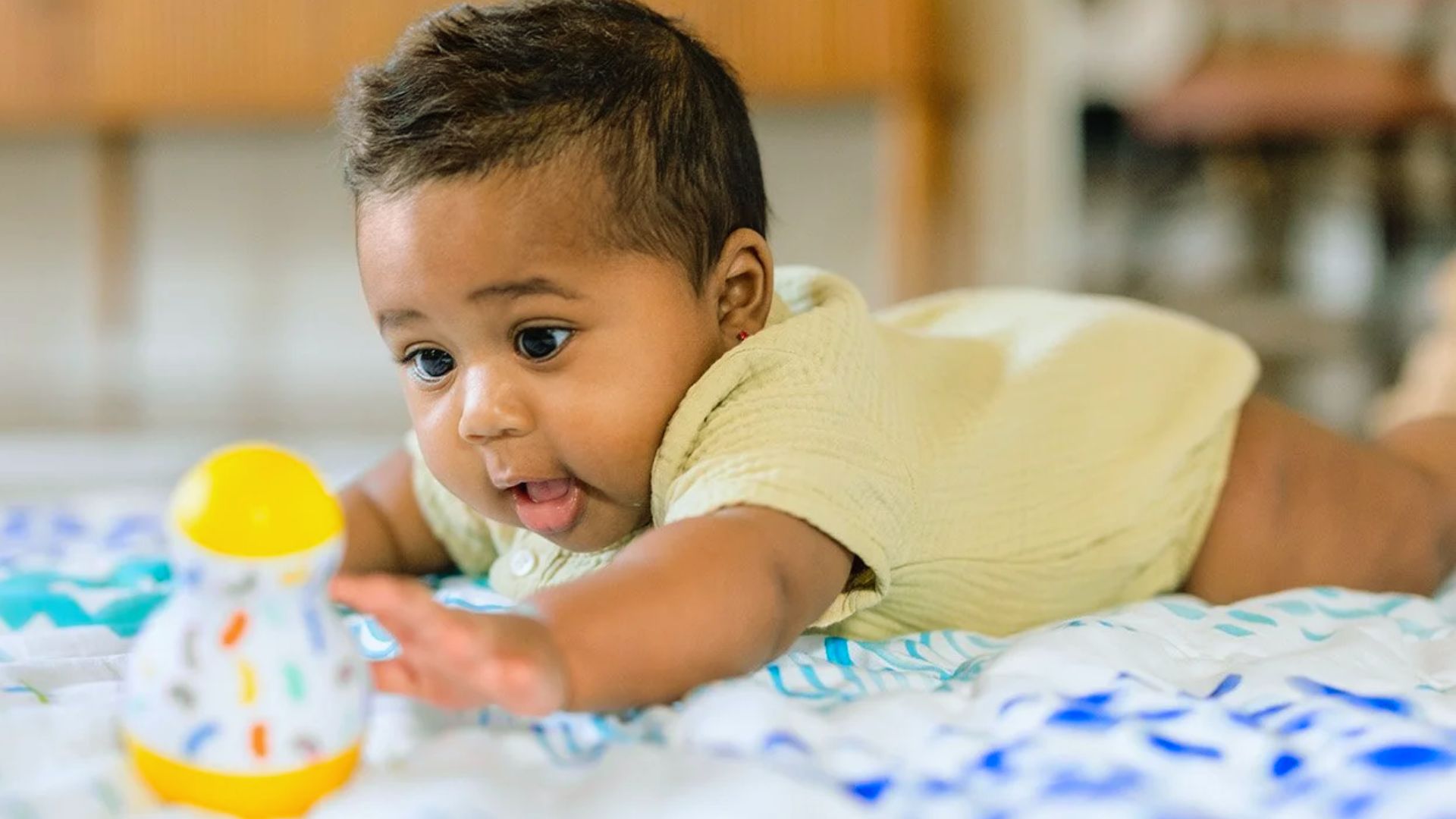 Baby in yellow outfit lying on patterned blanket reaching for colorful rattle toy