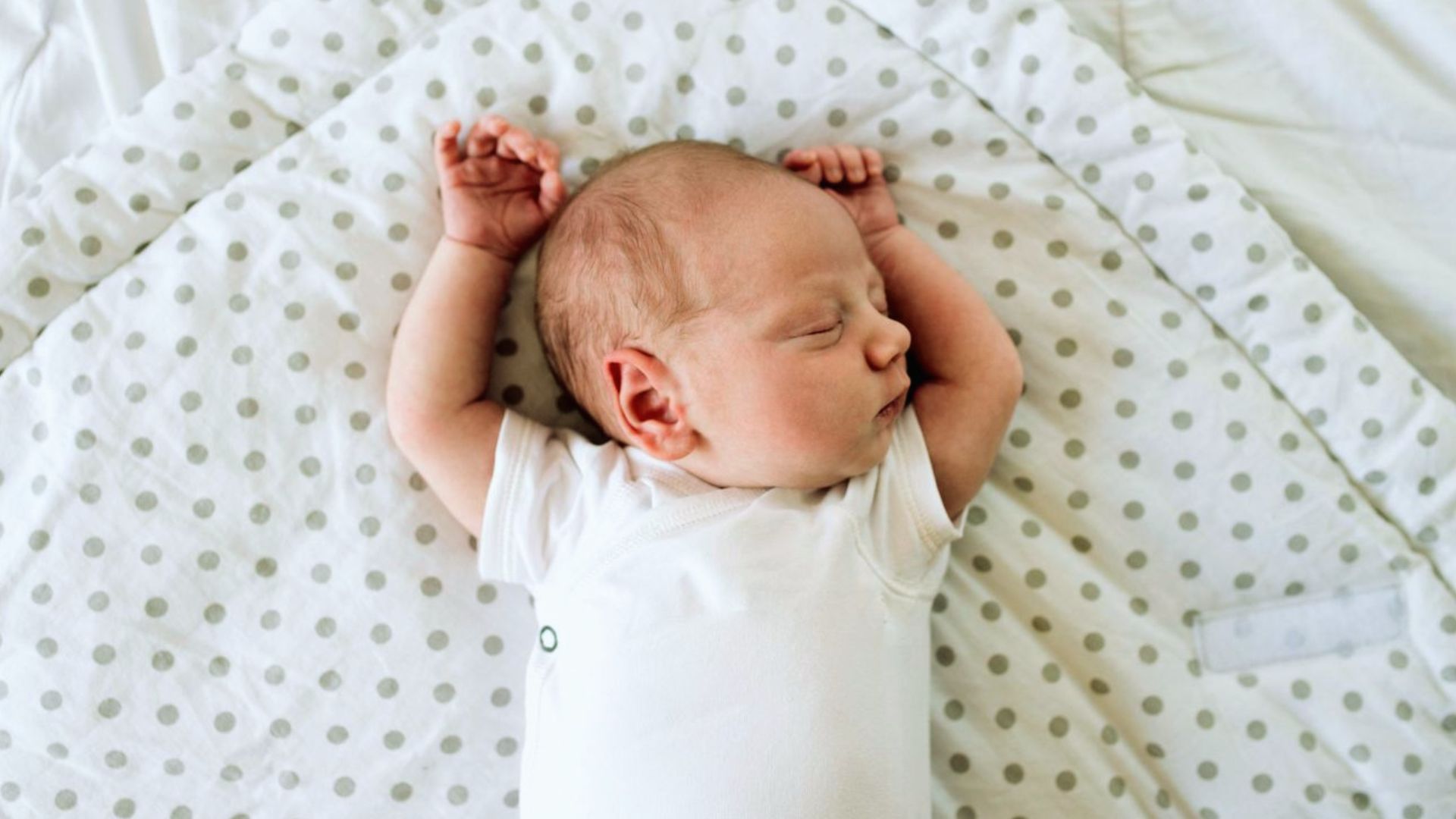 Newborn baby sleeping on polka-dot blanket wearing white onesie with arms raised