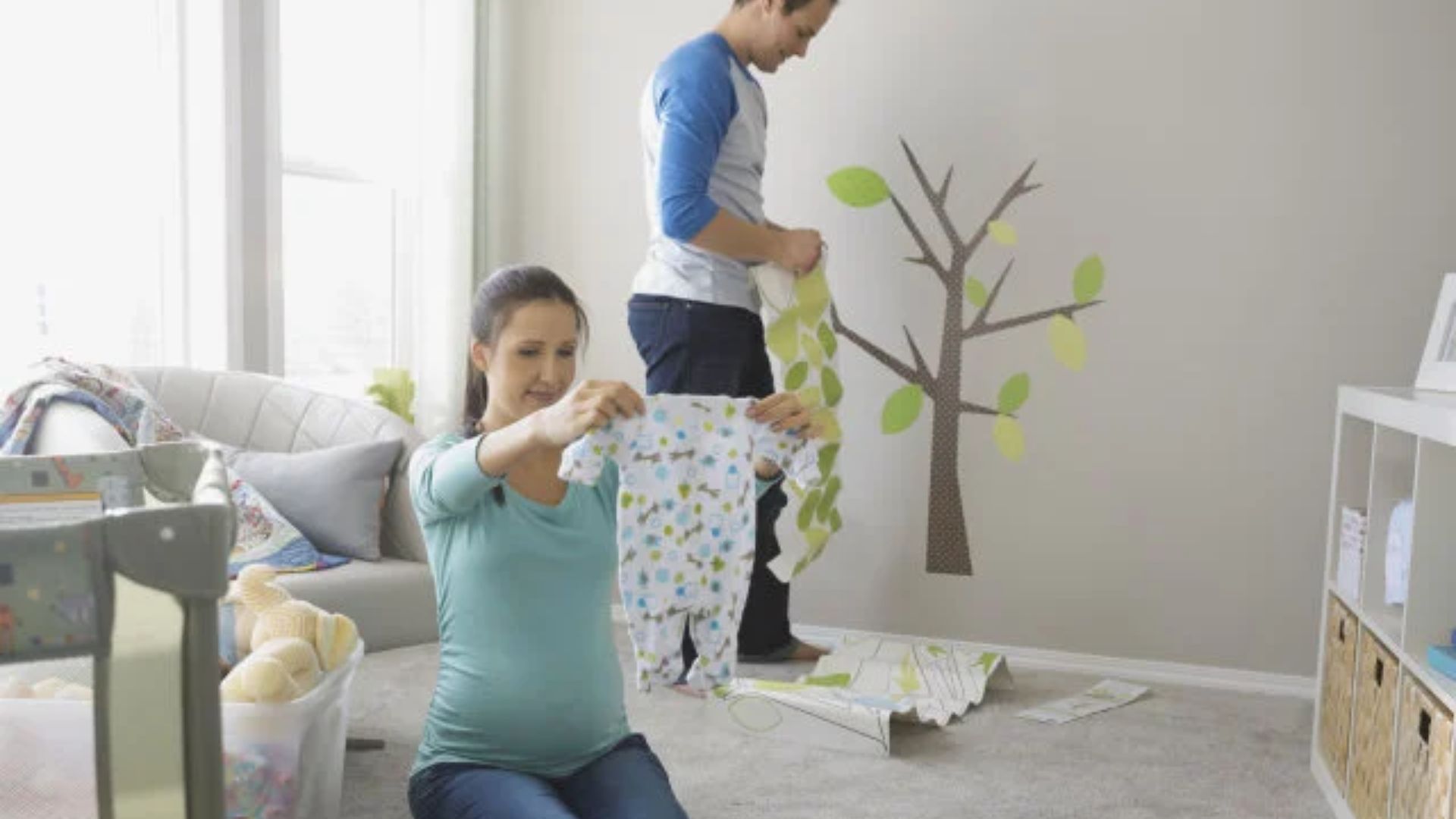 Pregnant woman kneeling holding up baby onesie with colorful print, man behind holding green leaf wallpaper in nursery with tree decal.