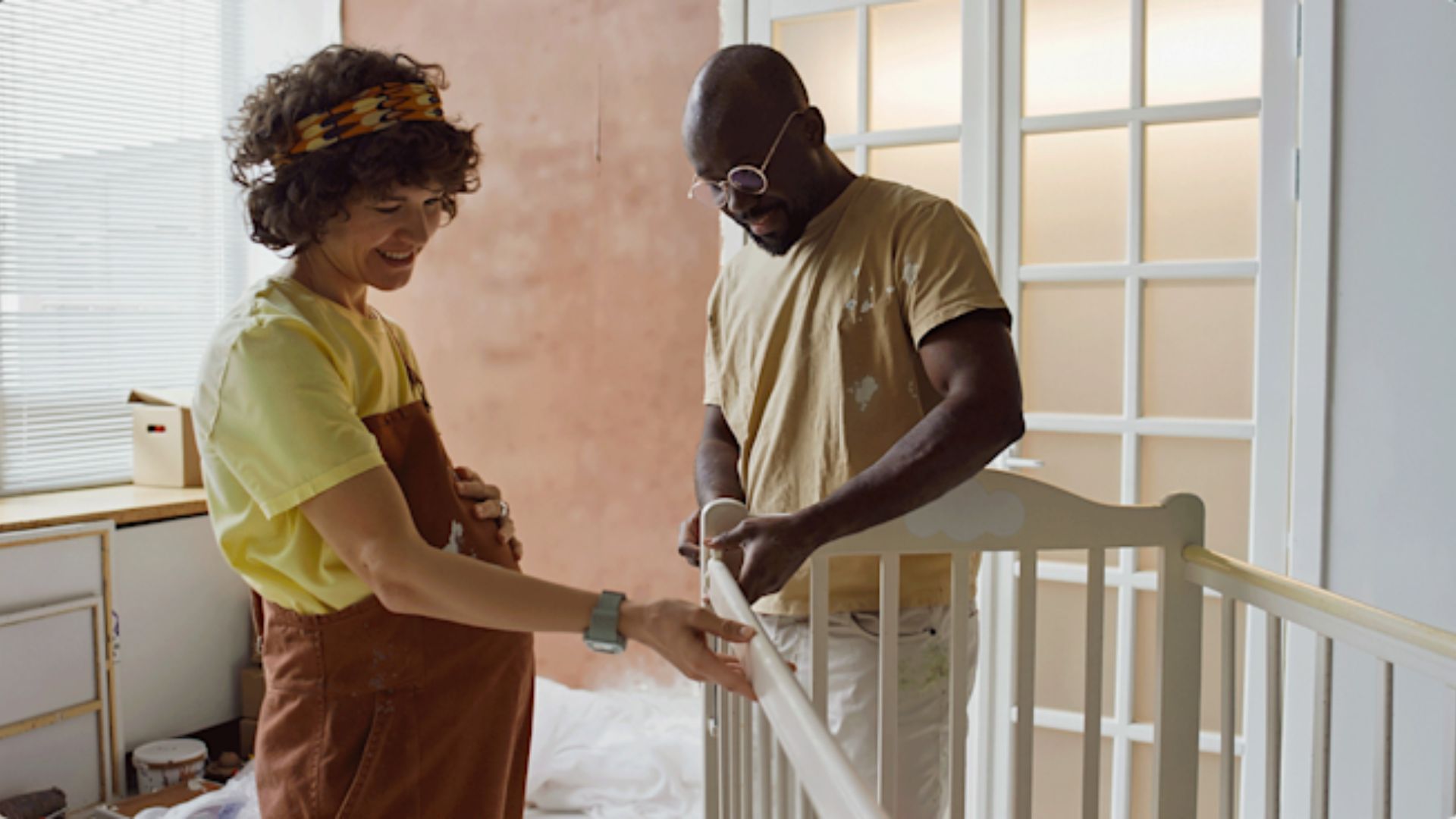 Pregnant woman in brown overalls and man in paint-splattered shirt assembling white baby crib together during room renovation.