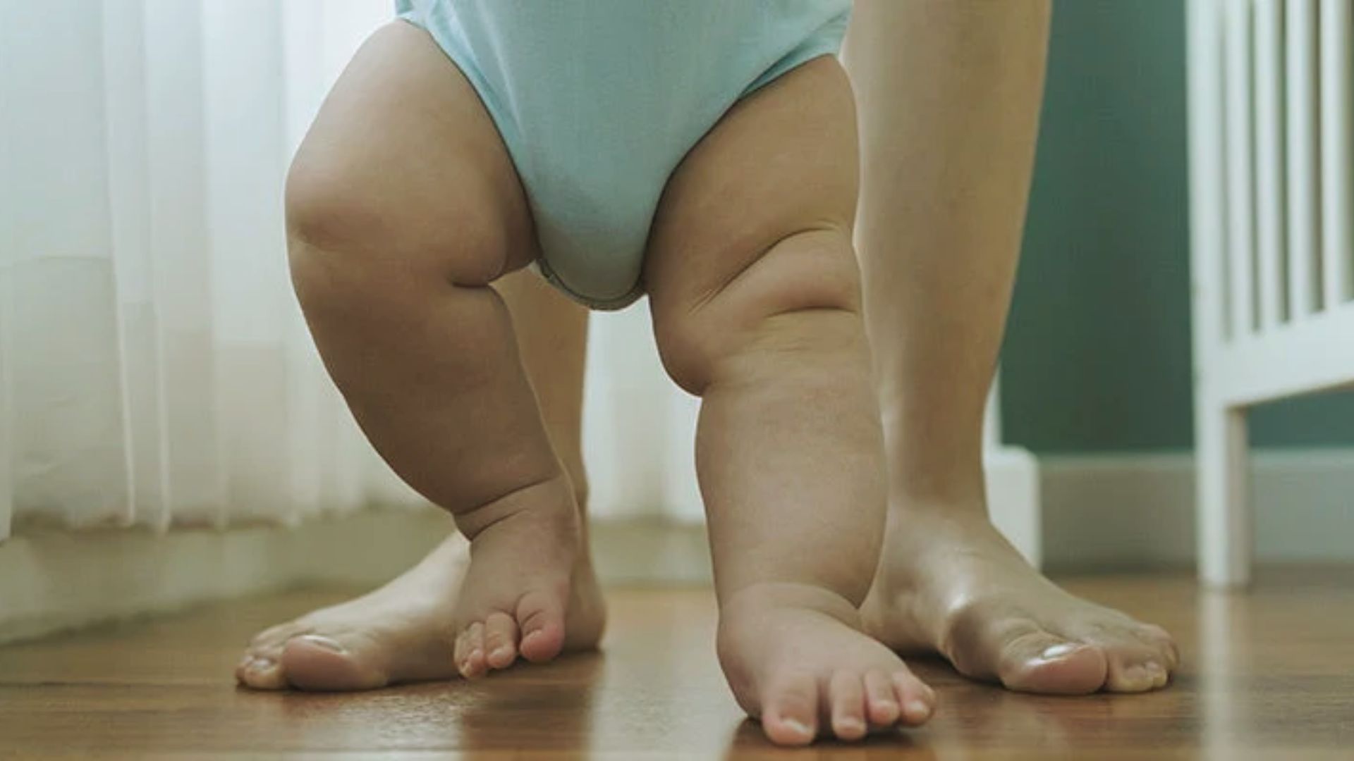 Baby's chubby legs taking first steps on wooden floor beside adult's bare feet