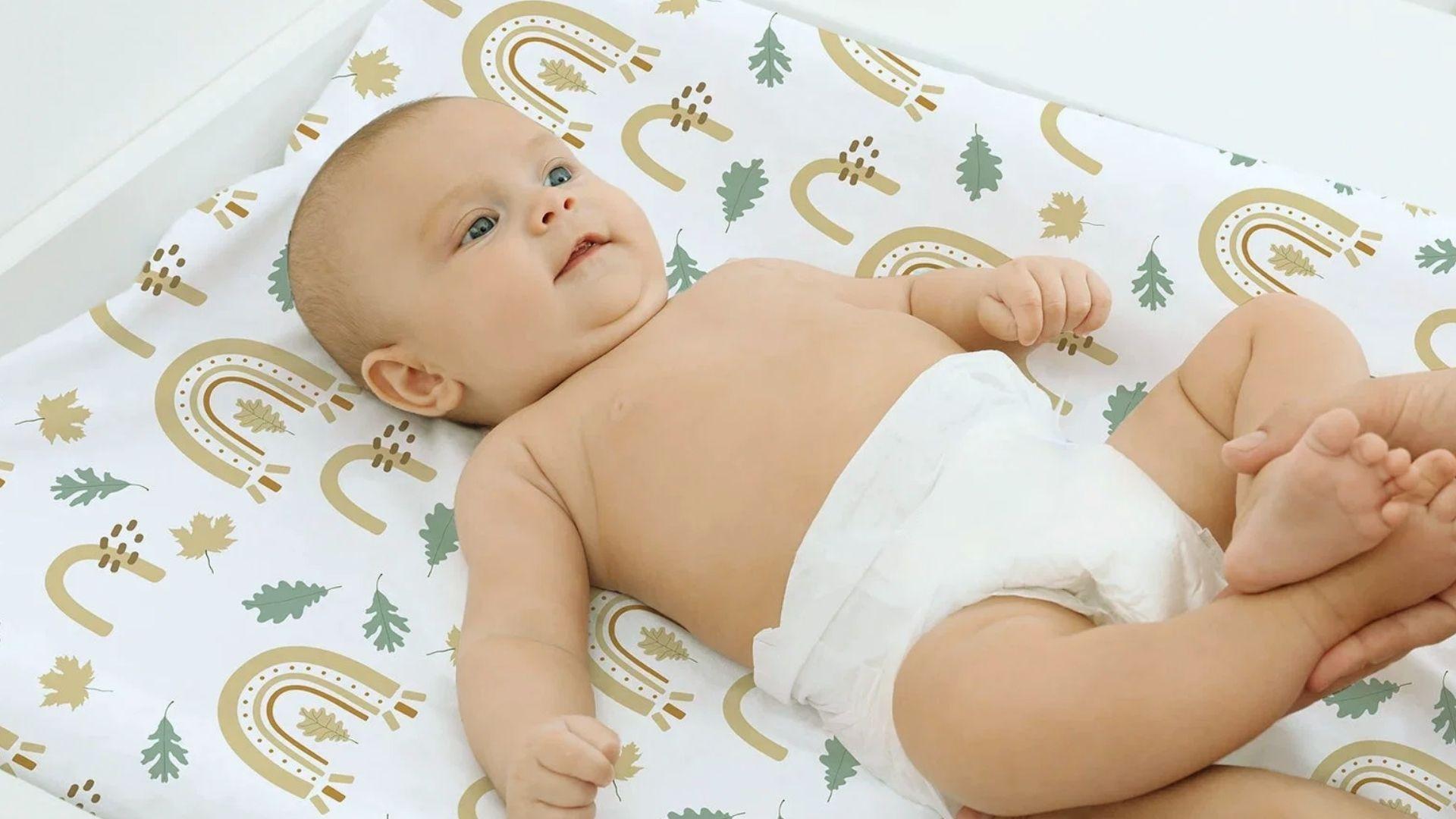 Baby in diaper lying in bassinet on rainbow leaf sheet, adult hands holding feet