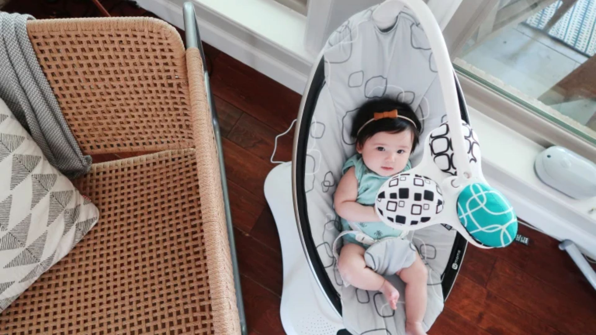 Baby with orange bow headband in black and white patterned mamaroo swing with hanging toys, beside wicker chair by window.