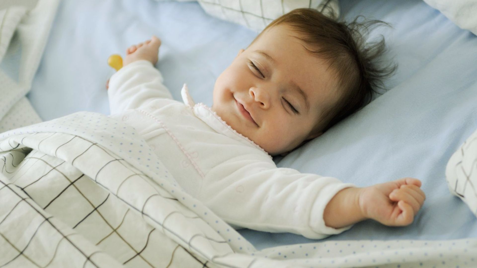 Smiling baby sleeping peacefully on blue bedding in white onesie with arms outstretched