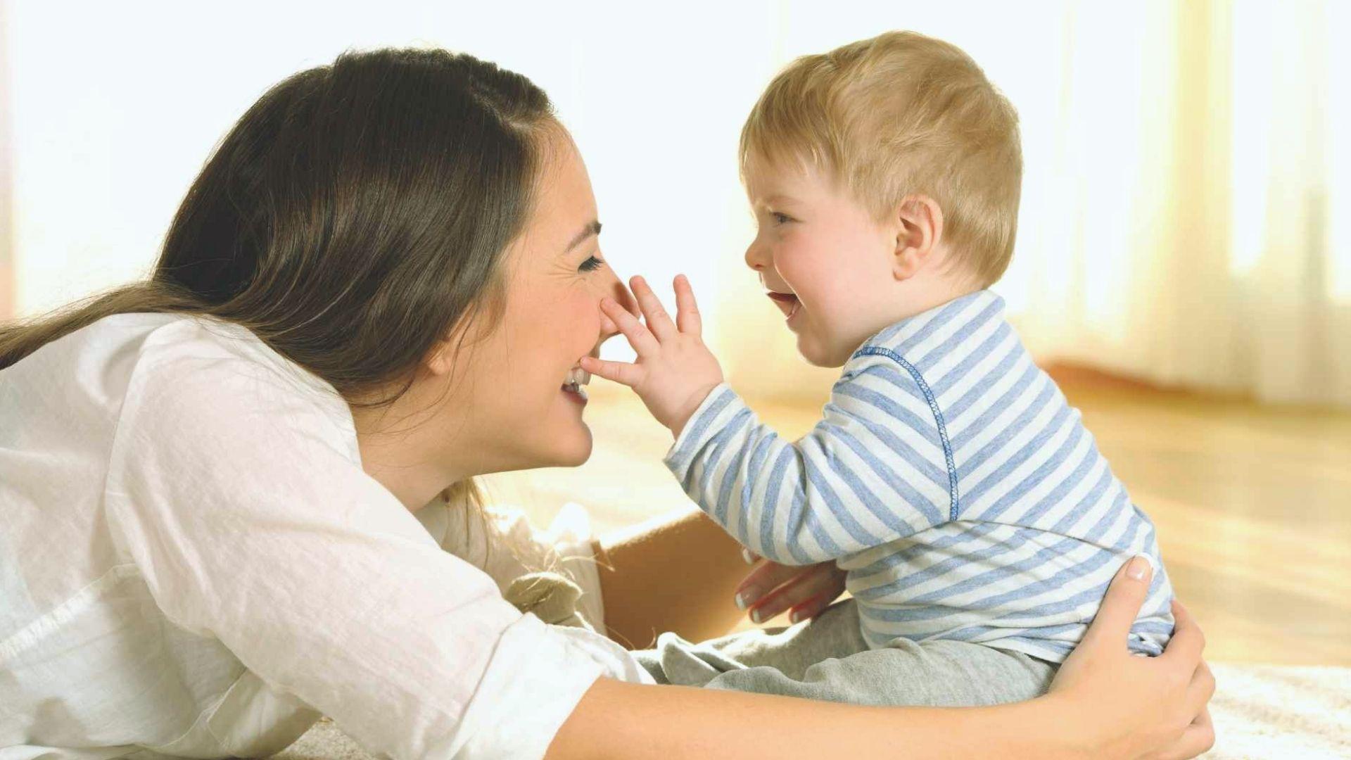 Happy mother laughing while baby in striped shirt playfully touches her nose indoors
