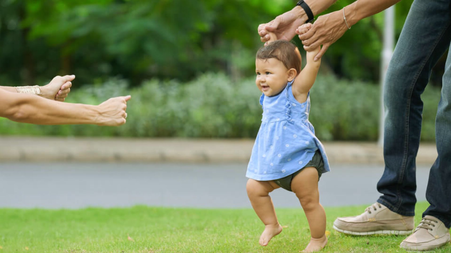 Baby in blue dress learning to walk on grass while held by adults reaching toward each other