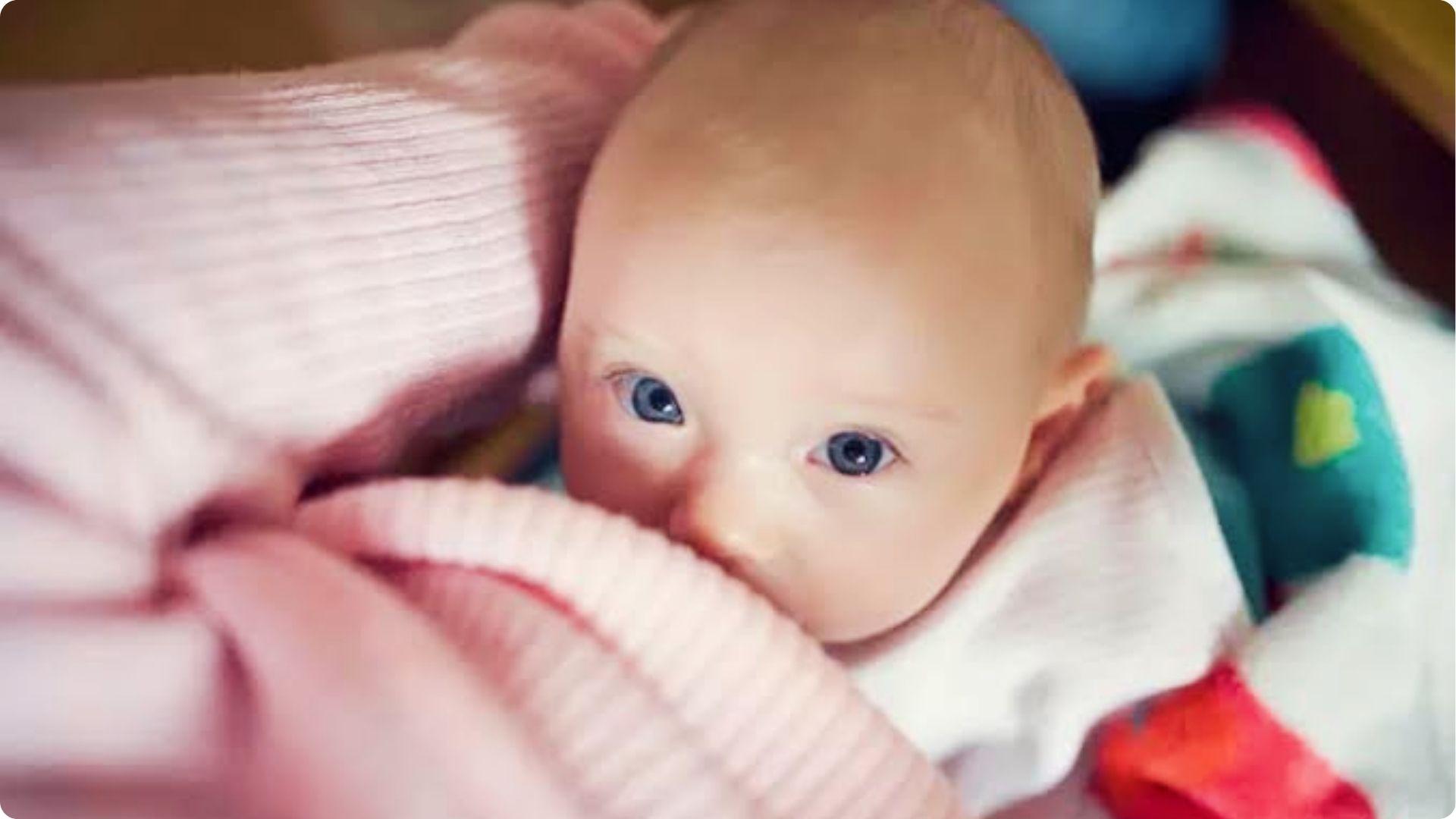 Blue-eyed baby peeking from a pink blanket, illustrating cozy warmth that makes babies start sweating.