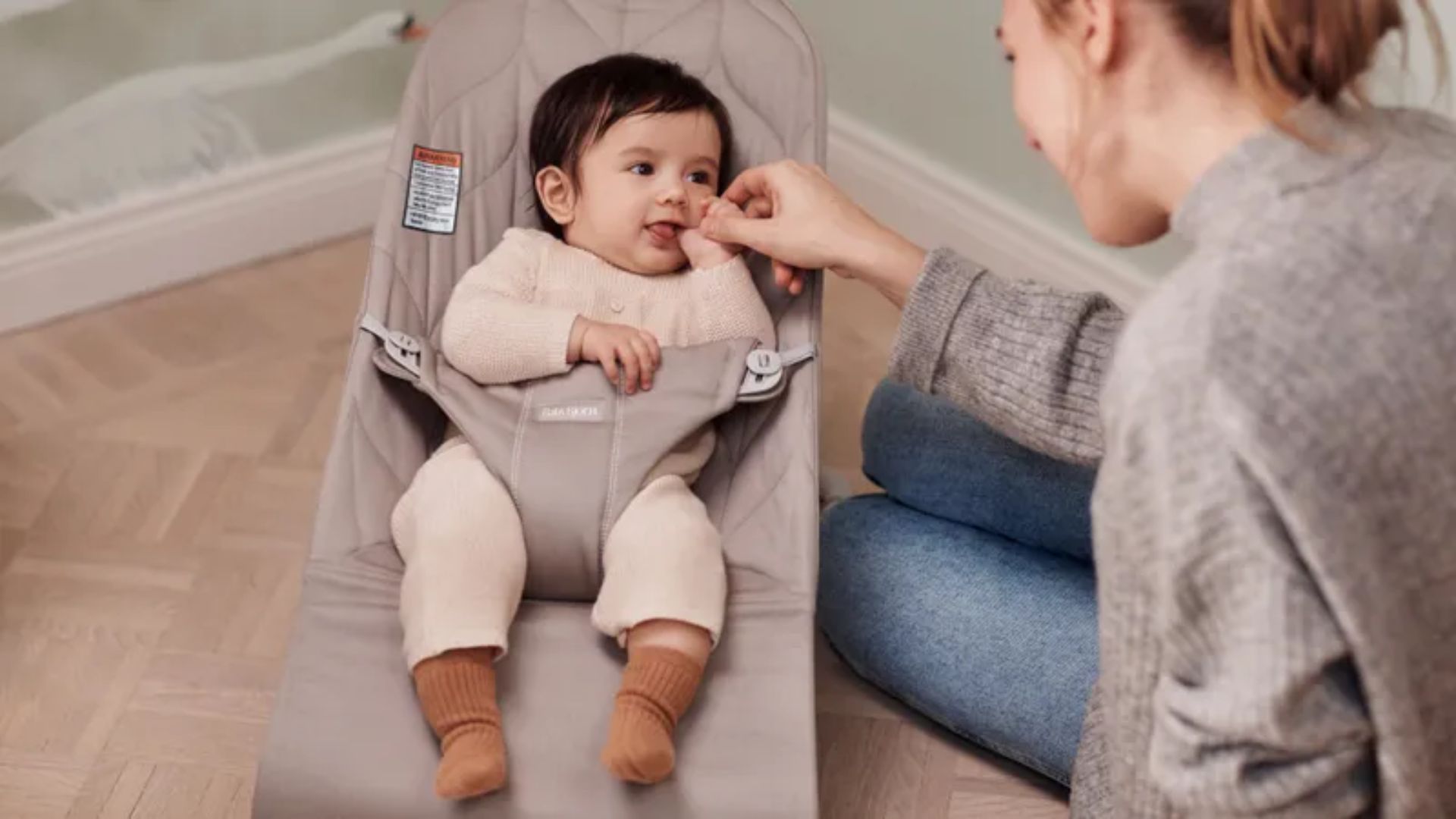 Baby in beige knit outfit with brown socks in taupe baby bouncer, mother in gray sweater kneeling and holding baby's hand.