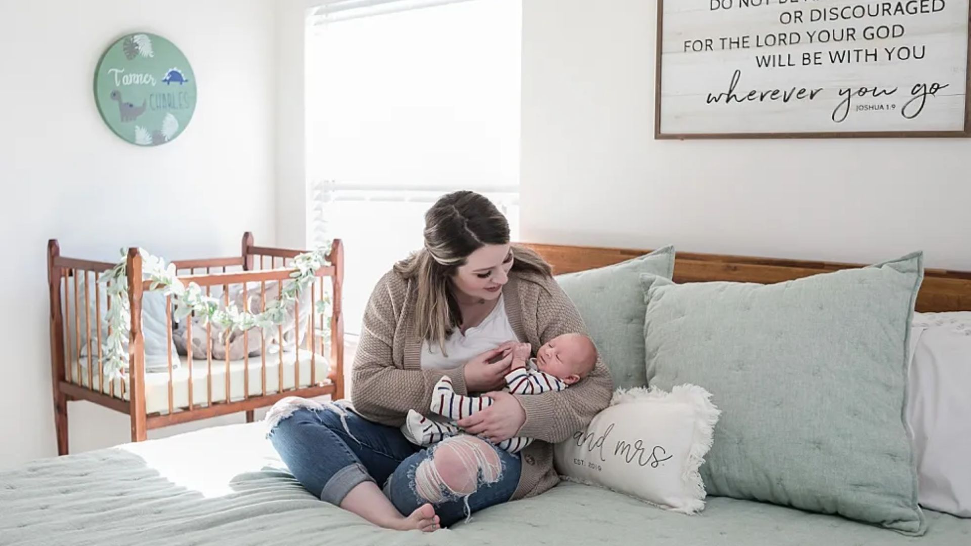 Mother in cardigan and ripped jeans sitting on bed holding newborn baby in striped onesie, wooden crib with garland behind.