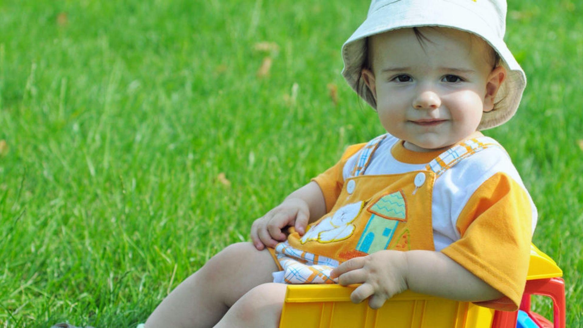 Cute baby in orange overalls and sun hat sitting in toy truck on green grass