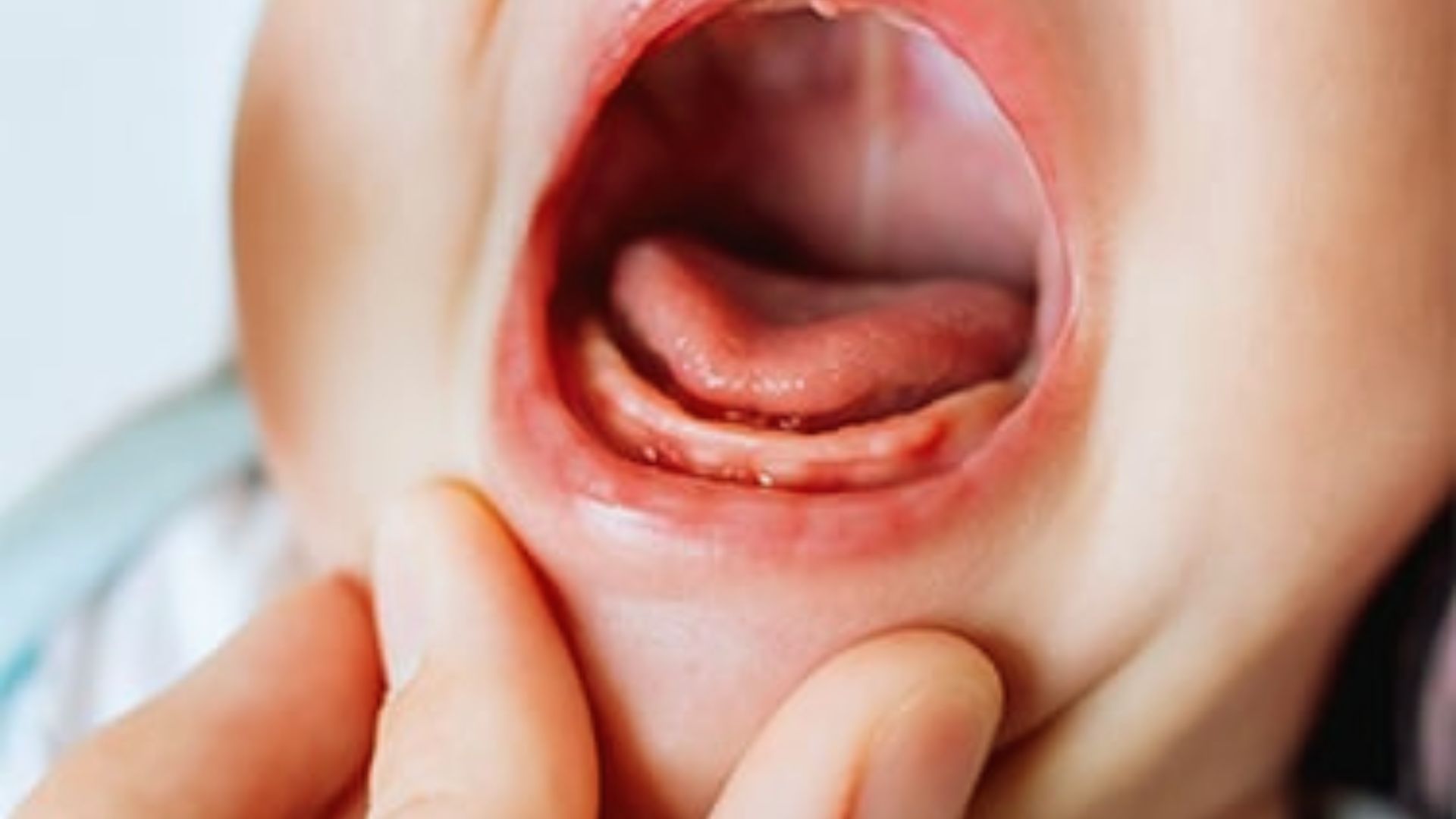 Close-up of baby with open mouth showing tongue and lower gums while adult fingers gently hold chin