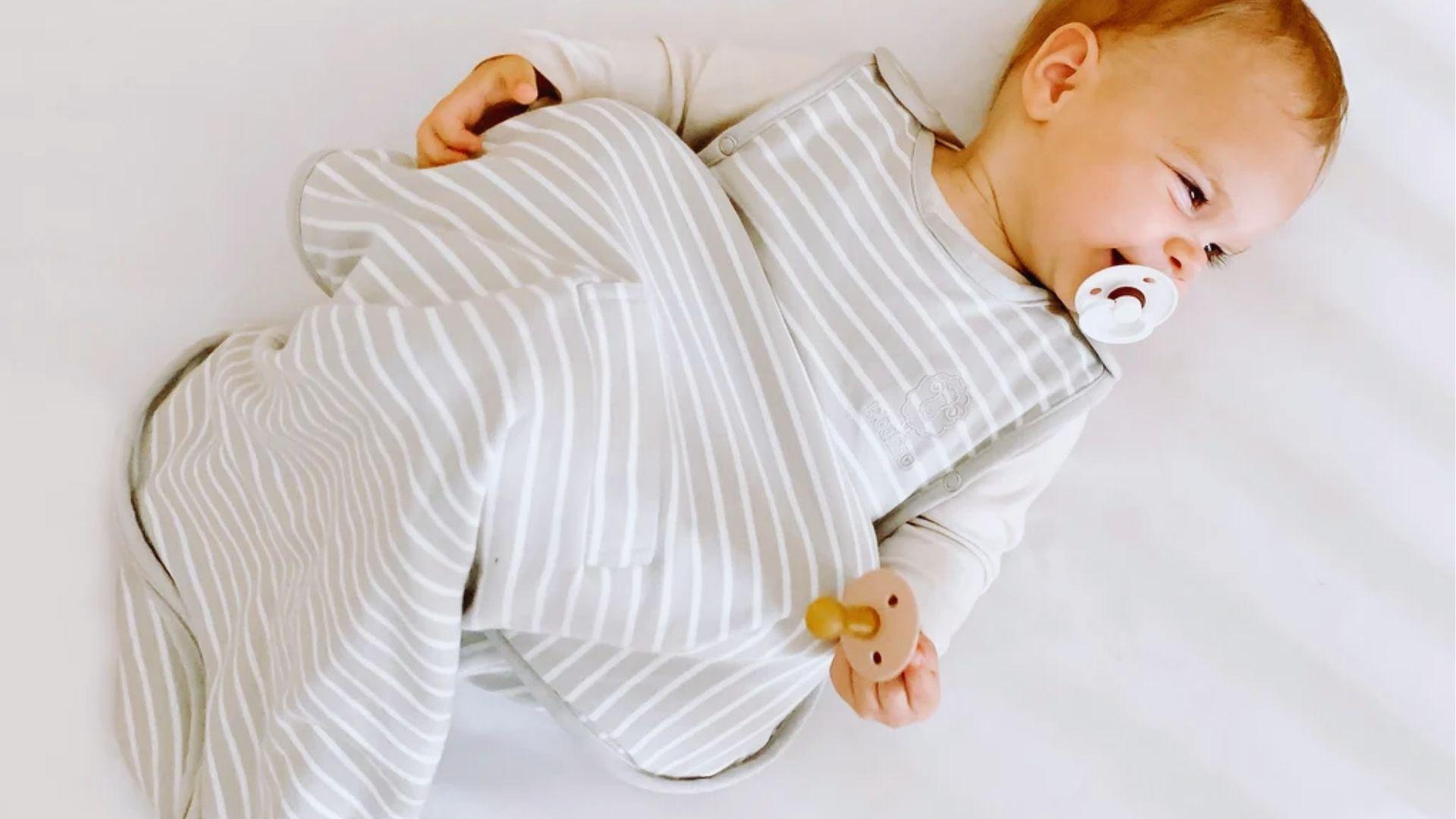 Woman in beige loungewear sitting on a light sofa smiling at a baby lying on a pillow, holding the baby's hand and foot