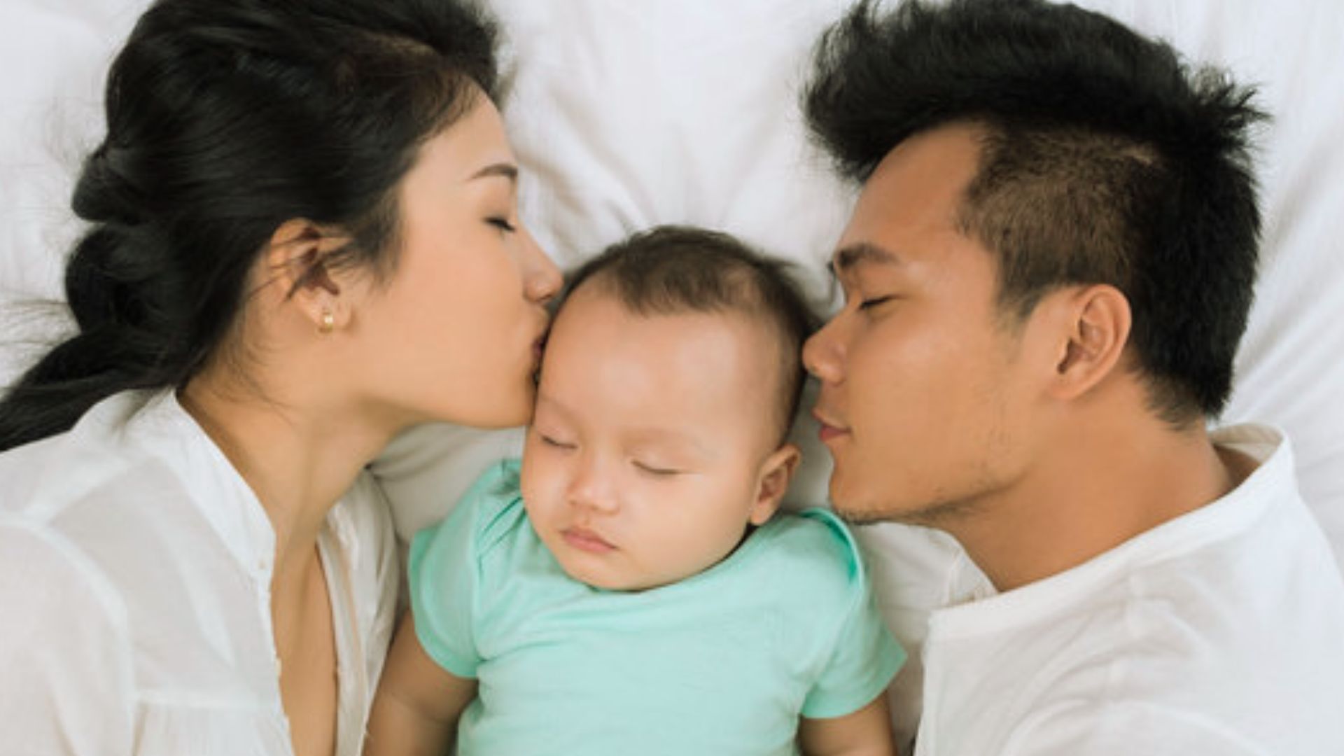 Parents lying in bed kissing sleeping baby in mint shirt between them