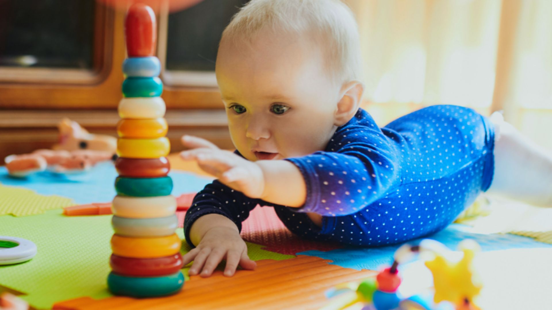 Baby in blue dotted outfit reaching for colorful stacking ring tower on play mat