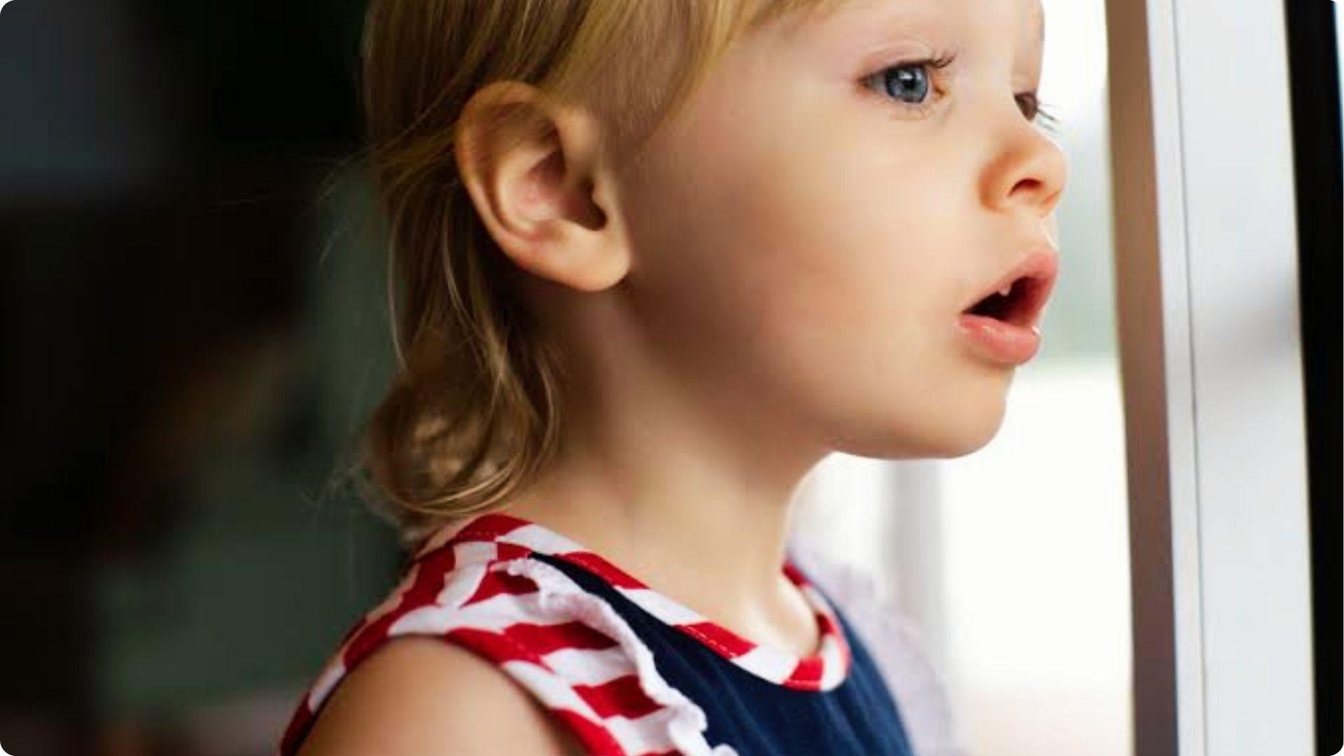 Little girl looks outside the window with open mouth in red and white striped top.
