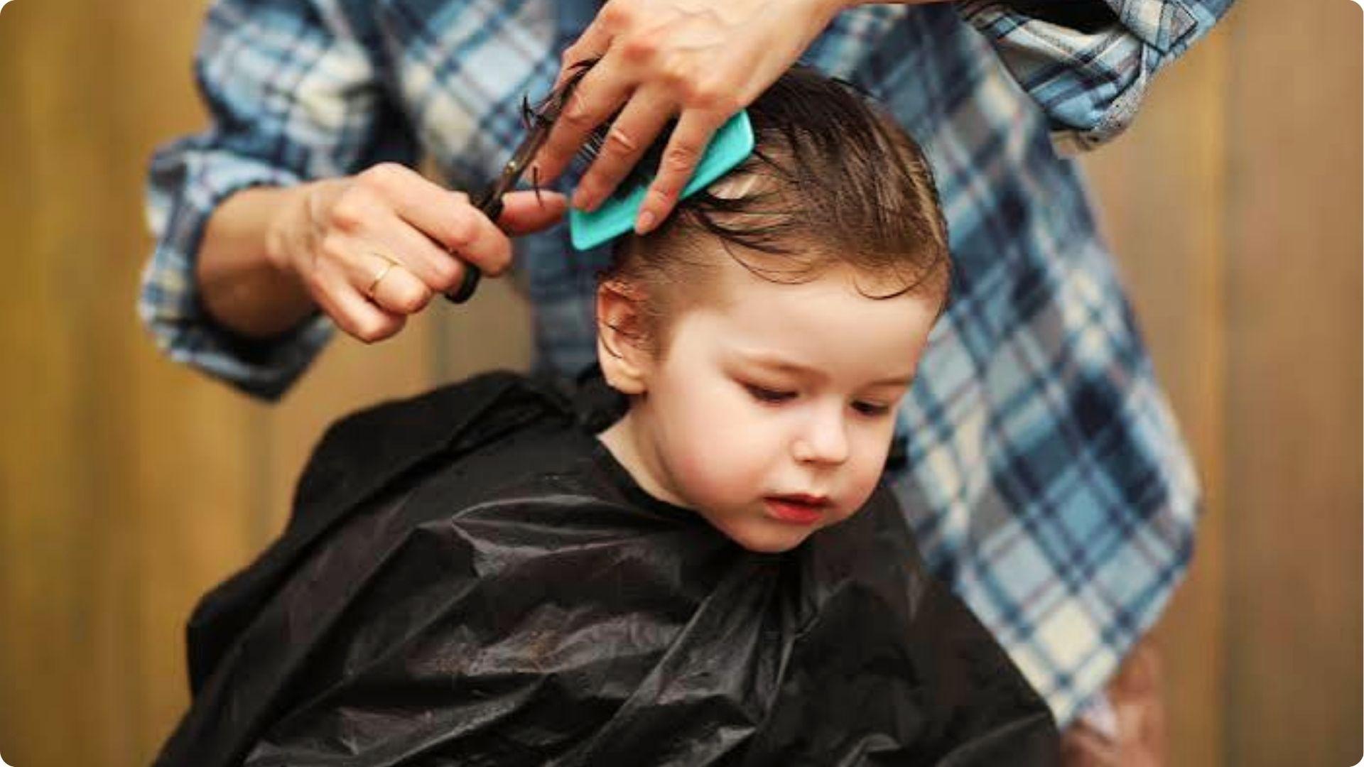 Toddler getting first haircut at home with scissors and blue comb, wearing black cape, cute trim.