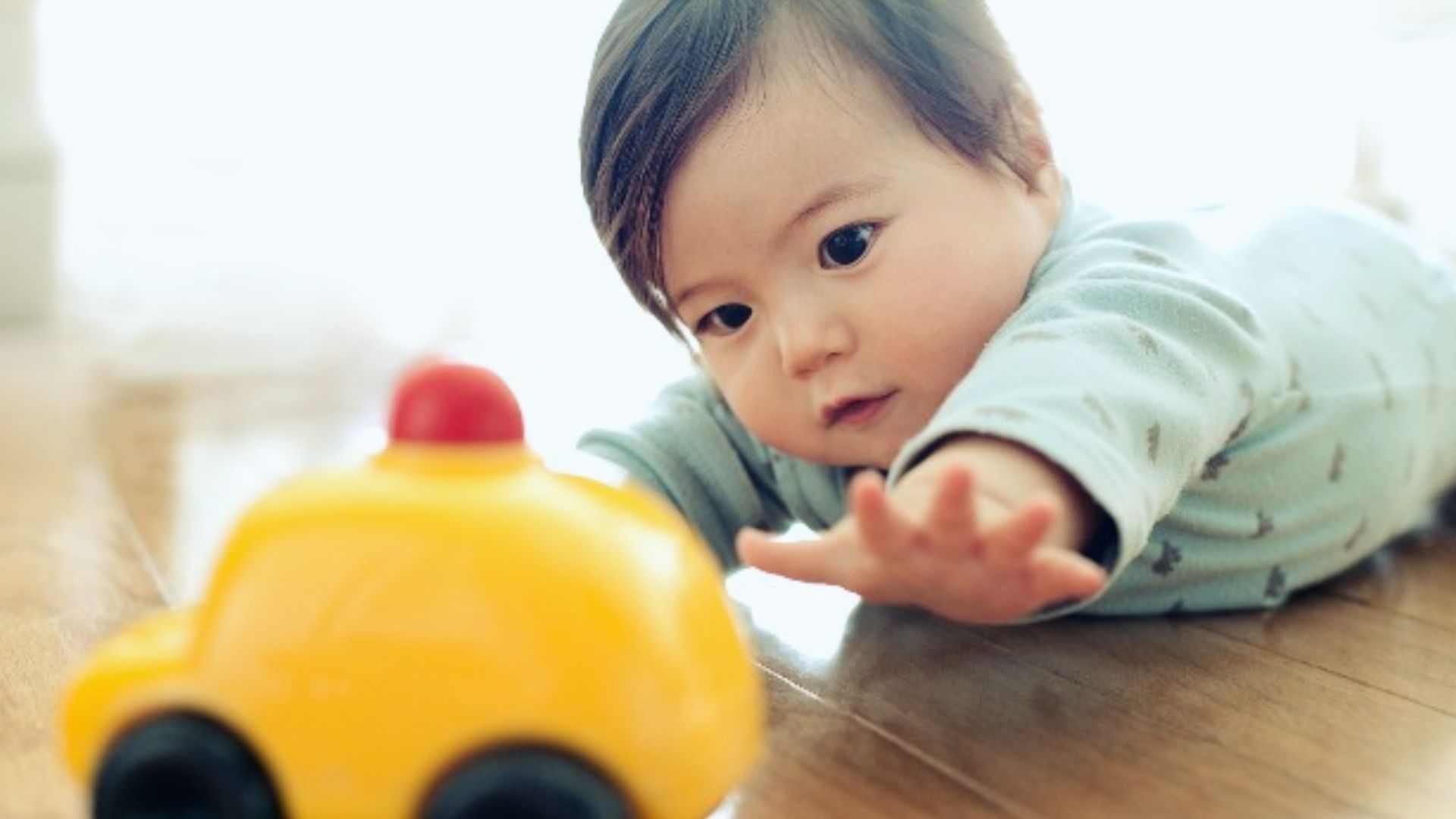Baby lying on wooden floor reaching for small yellow toy car