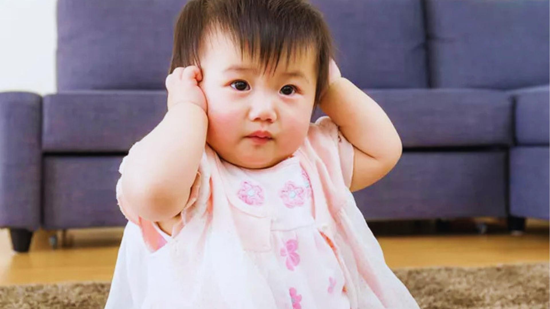 Toddler girl in pink dress holding both ears with hands standing in the living room today.