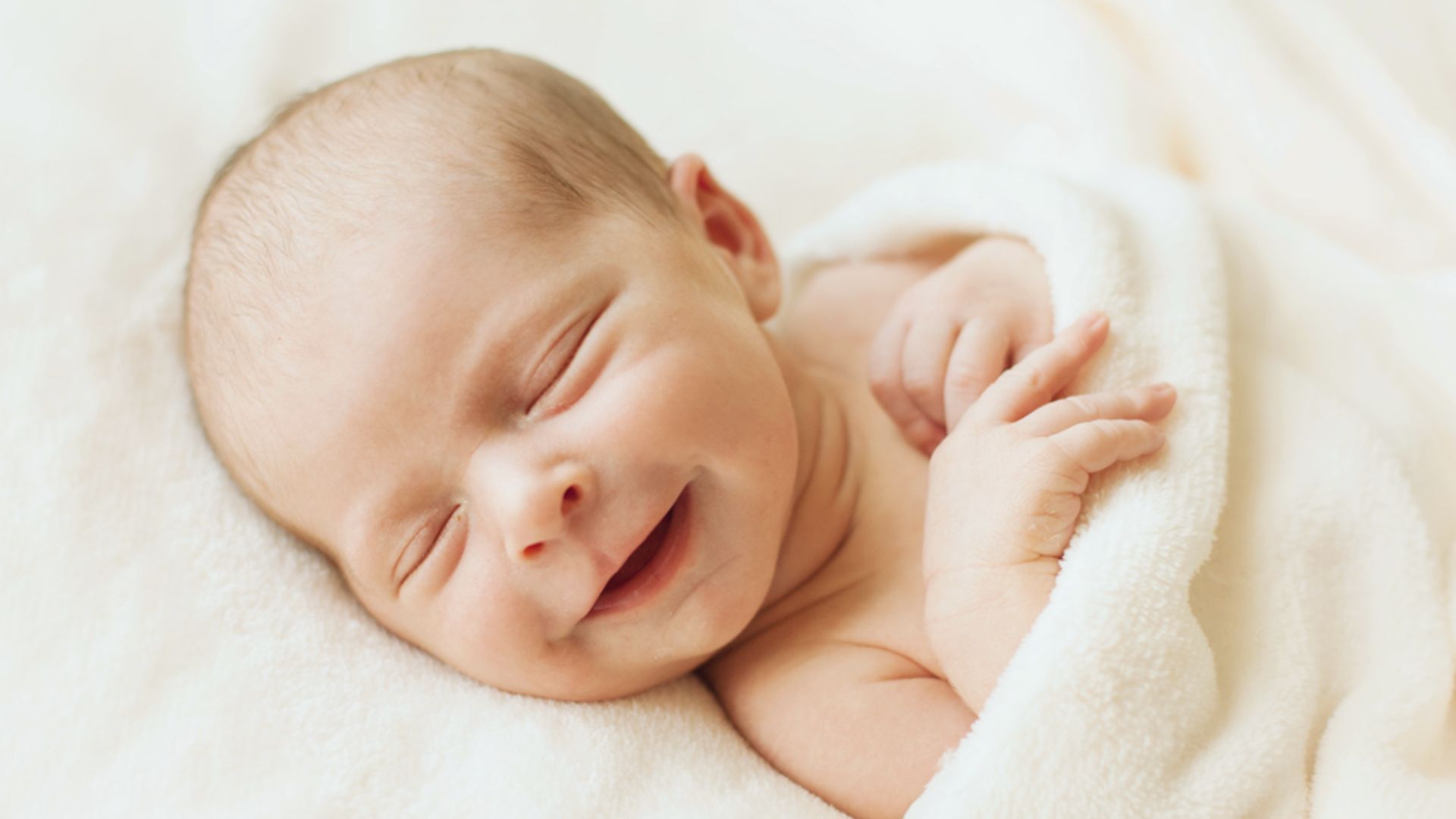Newborn baby smiling in sleep while lying swaddled in soft white blanket
