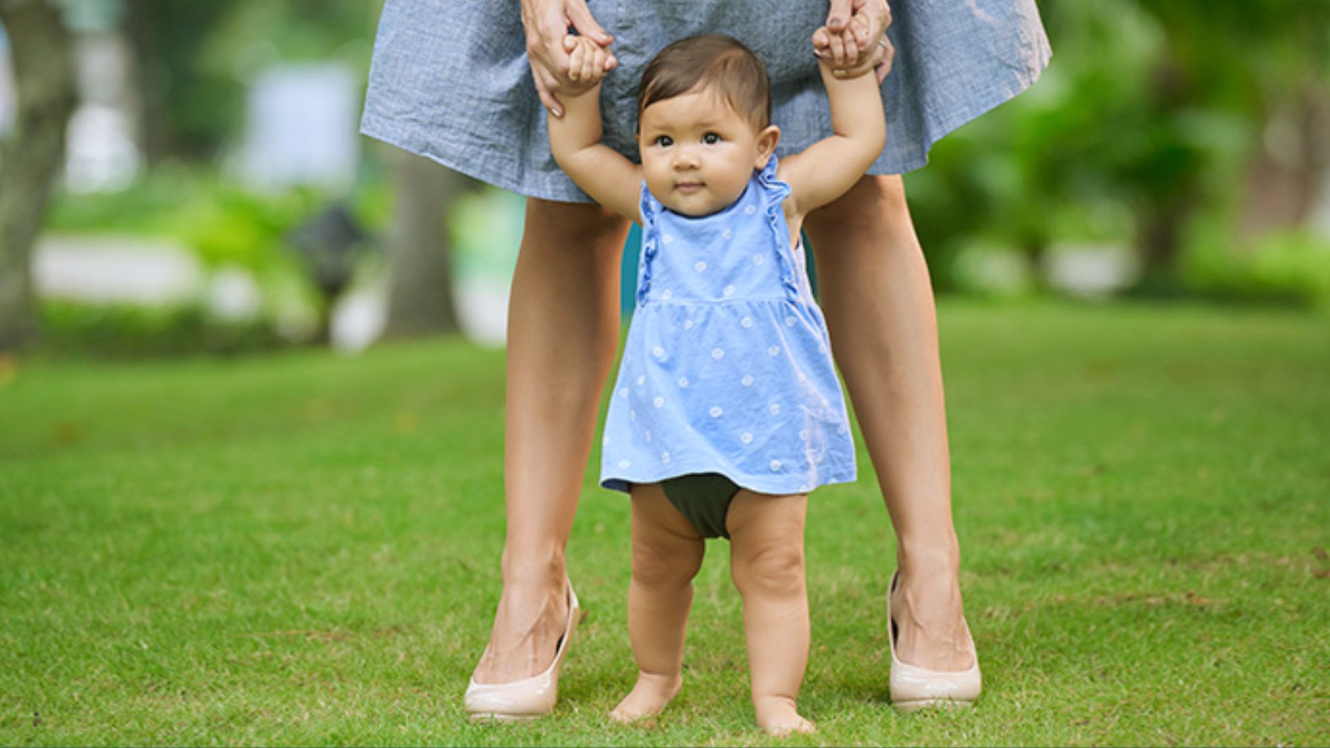 Toddler in blue polka-dot dress learning to walk on grass while holding adult's hands