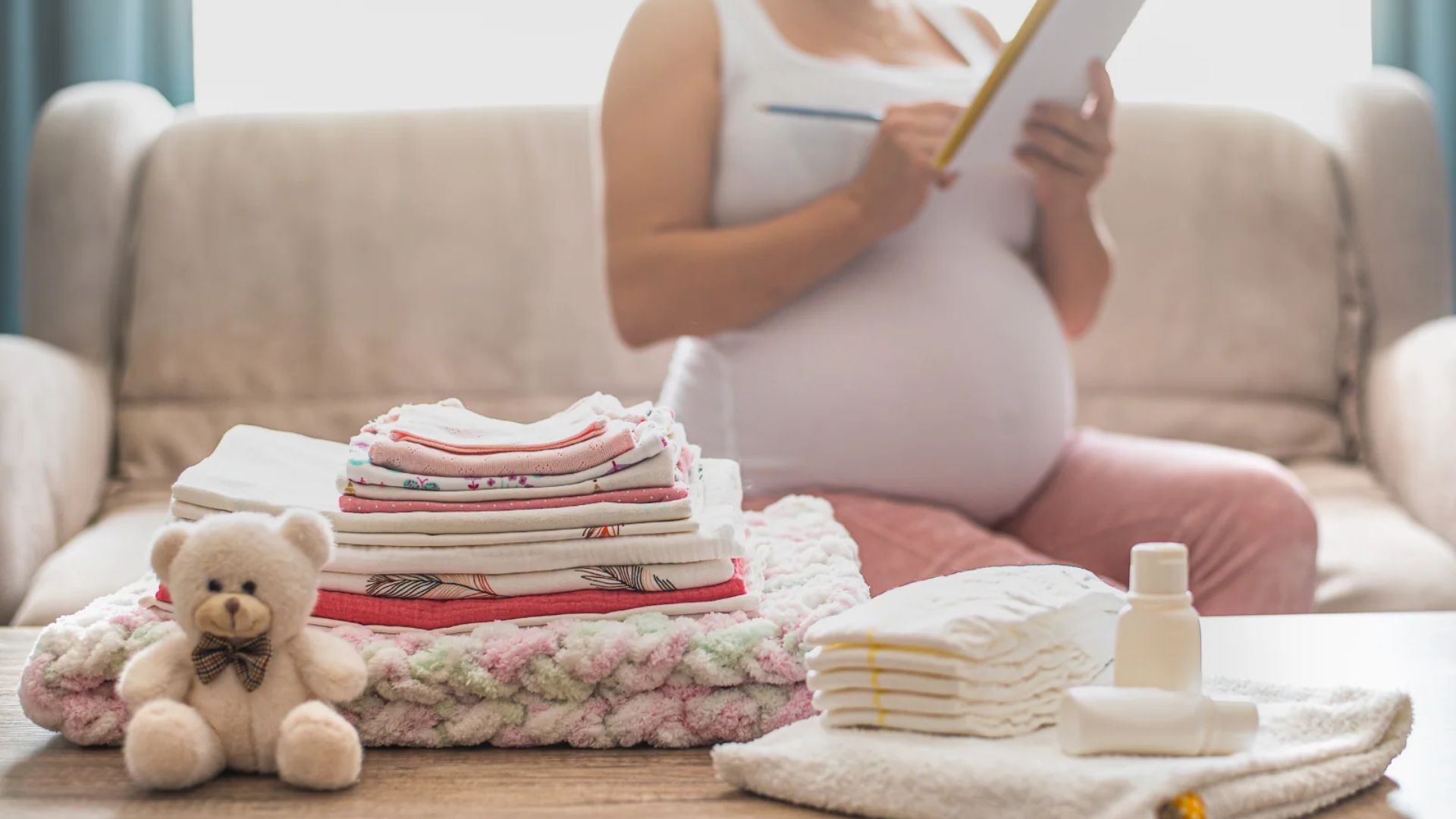 Stacked folded baby clothes and diapers with teddy bear on table in focus, pregnant woman in pink pants writing on notepad blurred behind.