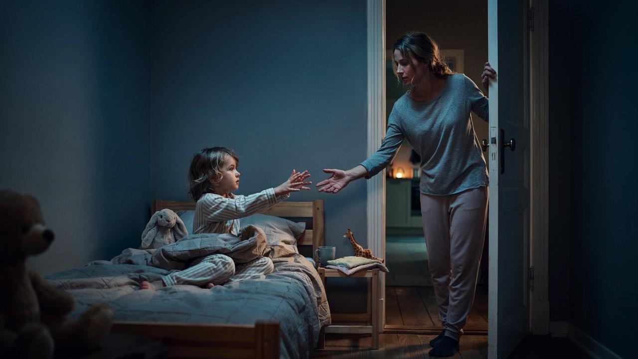 A toddler sitting up in bed at night reaching toward a parent standing in the doorway.