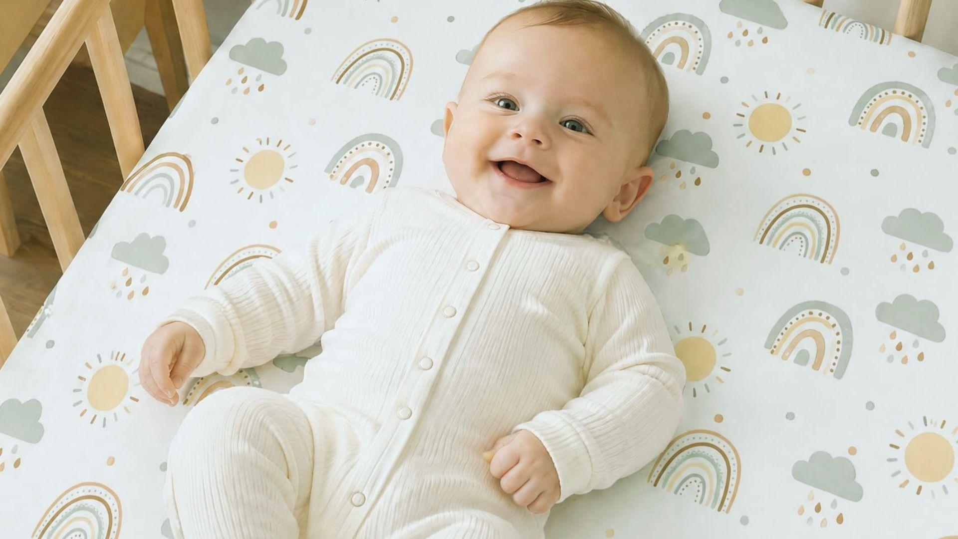 Smiling baby in white onesie lying in bassinet on rainbow sun cloud patterned sheet