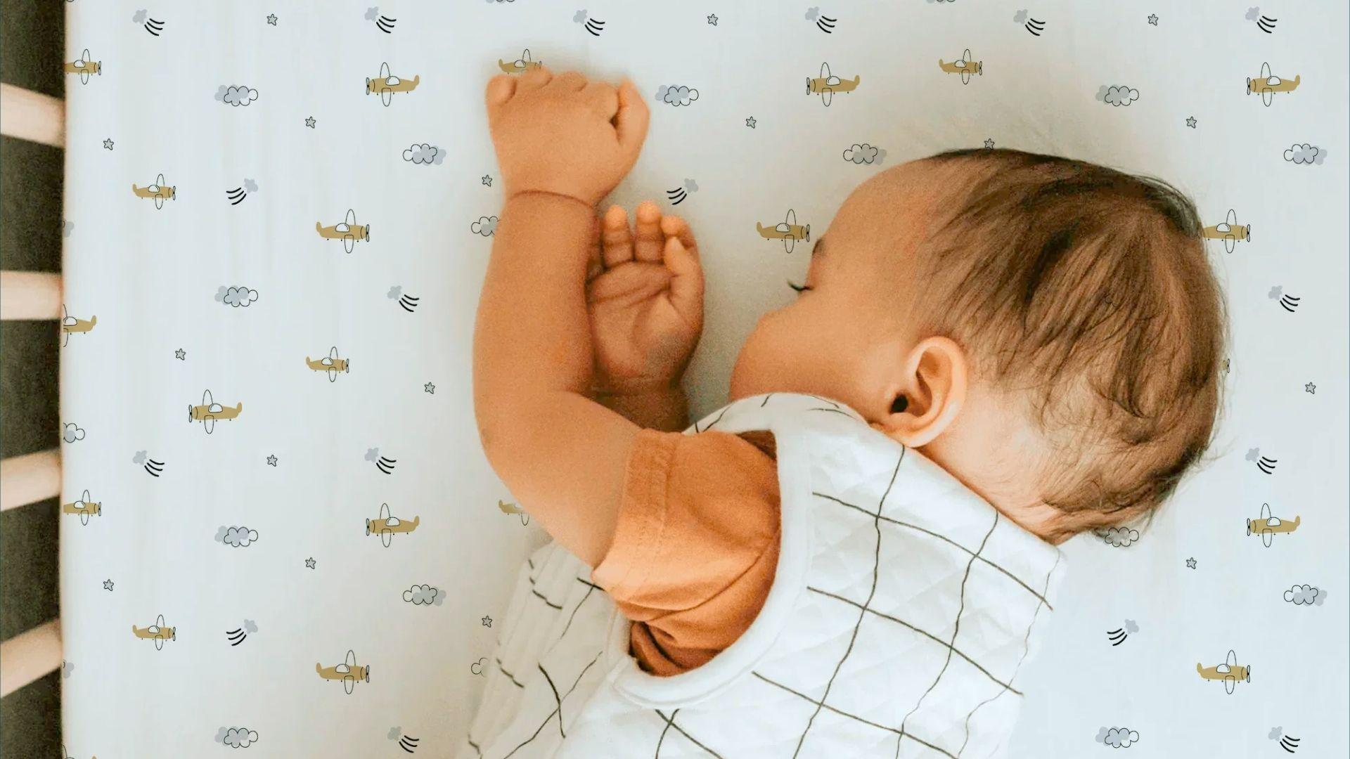 Baby sleeping in bassinet on airplane-pattern sheet, arms raised wearing checkered sleep sack