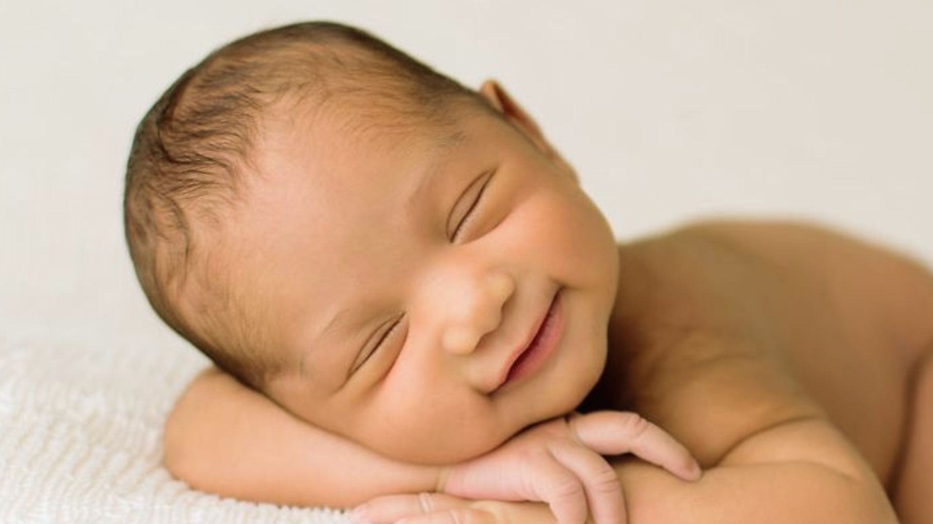 Newborn baby smiling in sleep while lying on white blanket with head on folded arms