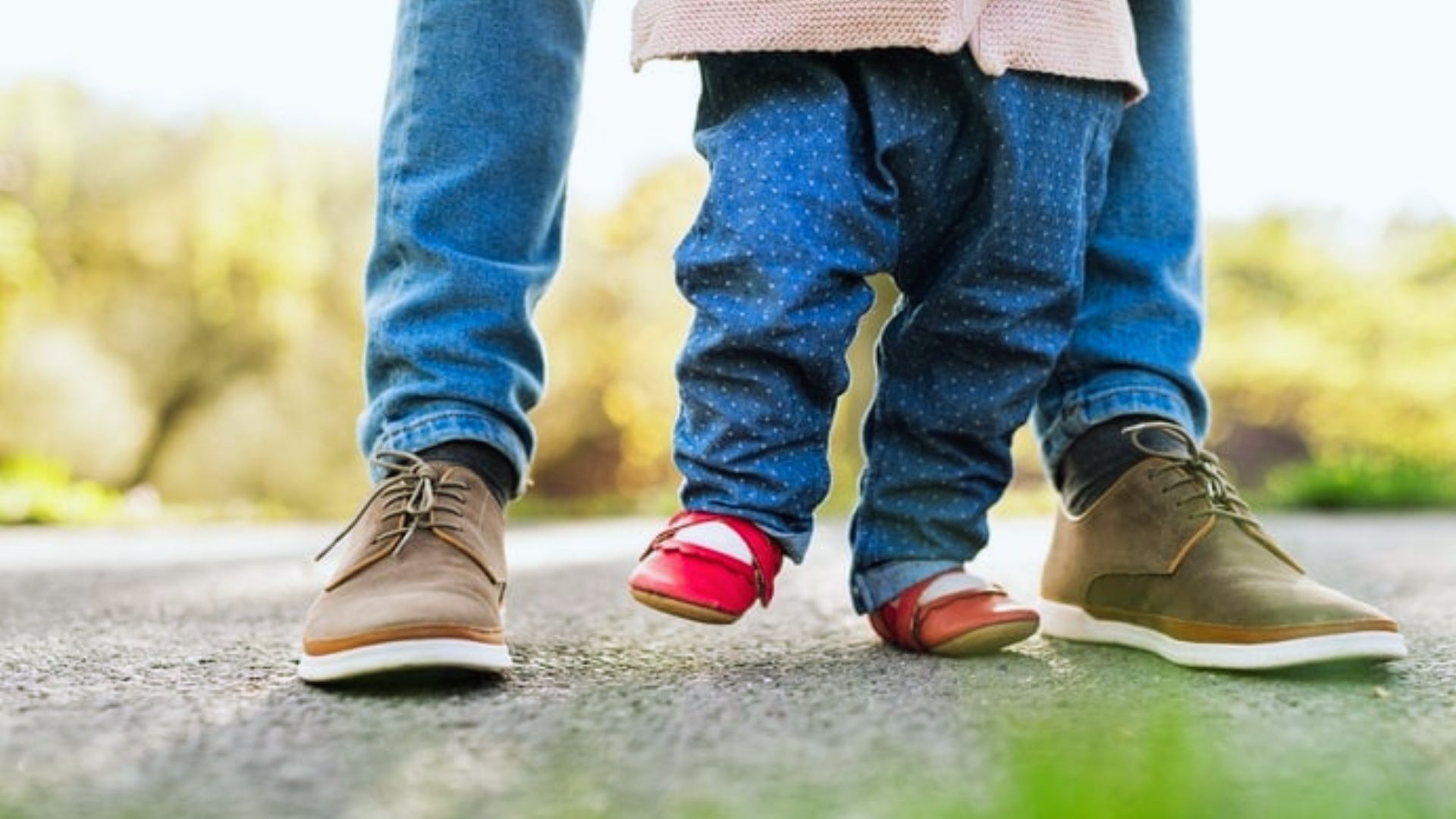 Toddler in red shoes stepping on adult's shoes while walking together on paved path outdoors
