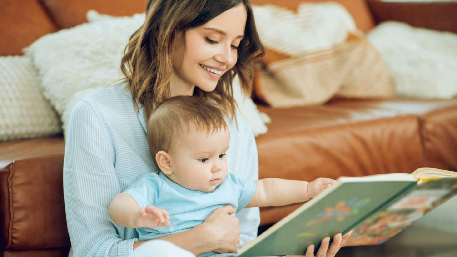 Smiling mother holding baby in blue shirt while reading colorful picture book together on couch