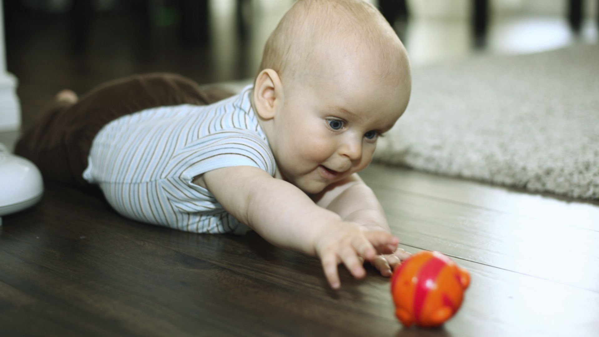 Baby lying on floor reaching for orange and pink striped ball toy