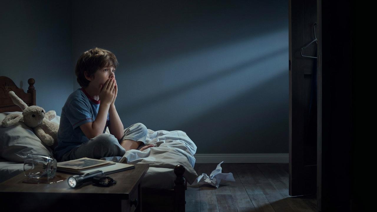 A young boy sitting up in a dark bedroom at night looking scared and worried.