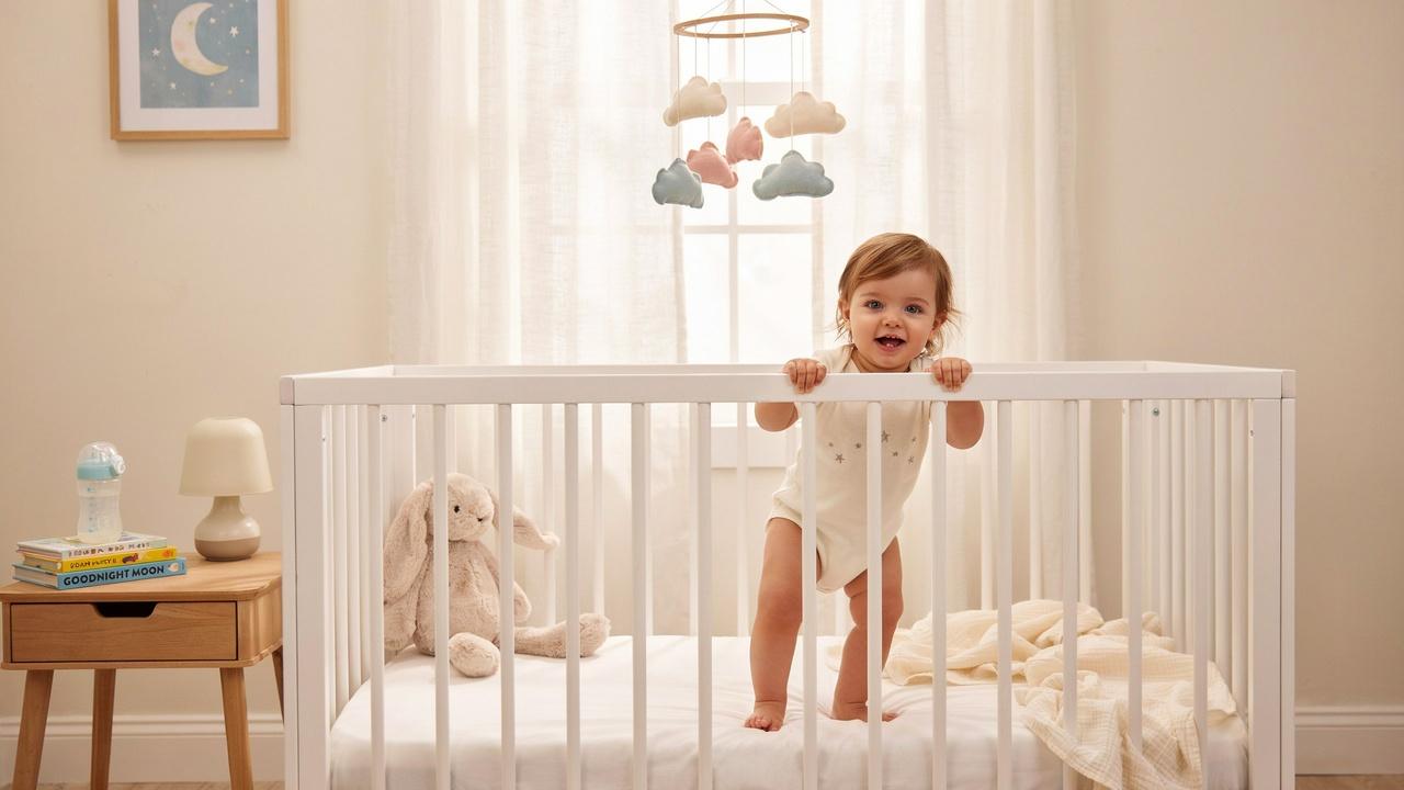 A smiling toddler standing up in a white crib during nap time.