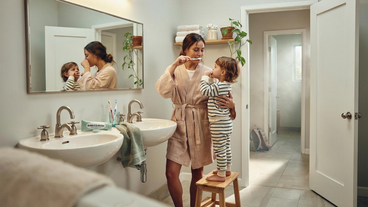 A parent and toddler brushing teeth together side by side in a clean bright bathroom.
