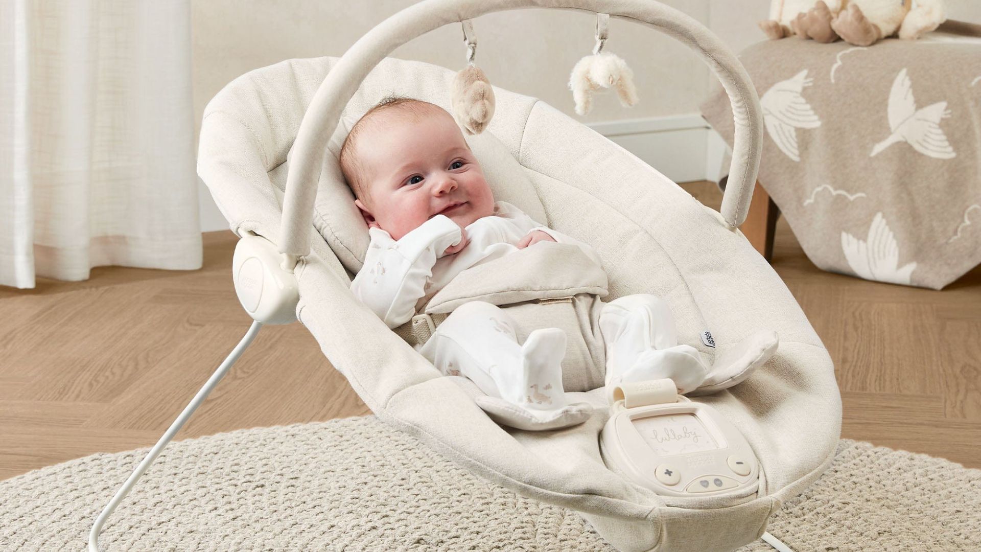 Smiling baby in white onesie in beige electric bouncer with plush toy bar and lullaby control panel, on woven rug.