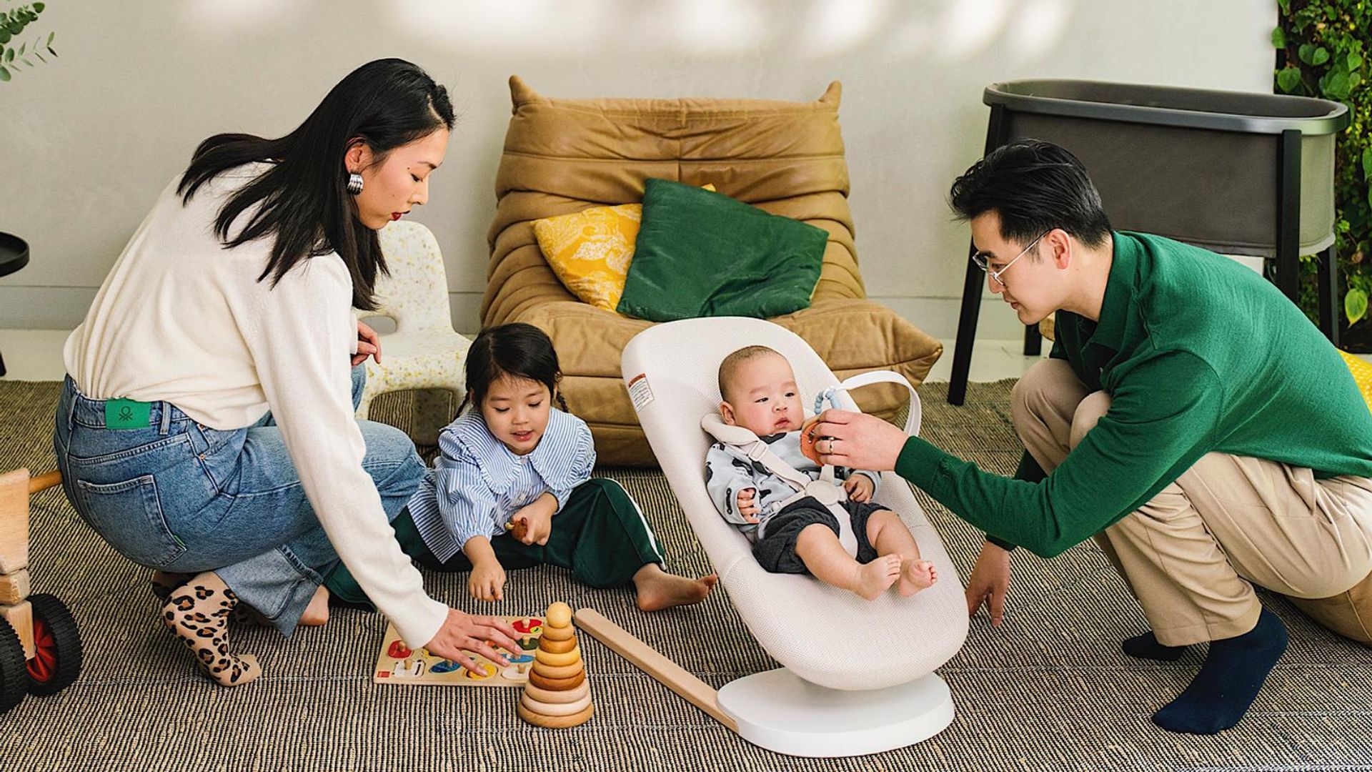 Asian parents playing on rug with toddler doing puzzle and baby in white bouncer, living room with brown chair and bassinet.