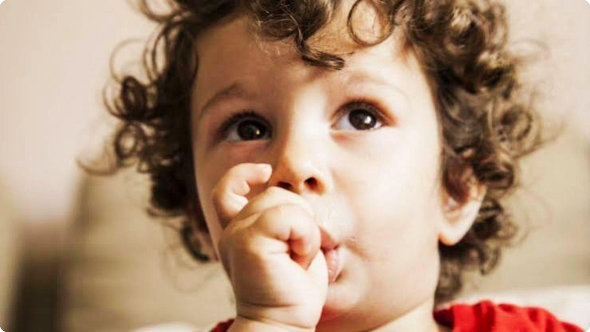 Curly haired toddler sucking thumb while looking up, common self comfort behavior from newborn months