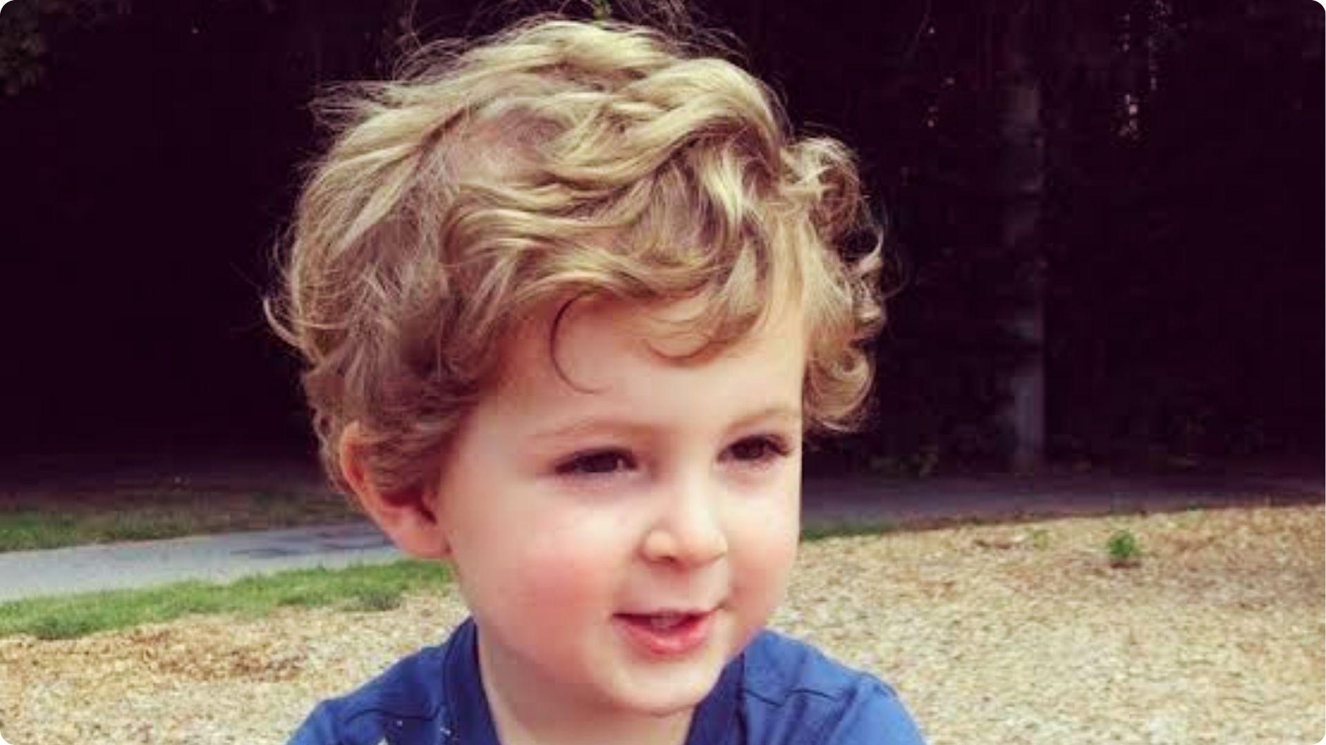 Blonde toddler with wavy curly top and side-swept curls, outdoor light, cute baby boy haircut style.