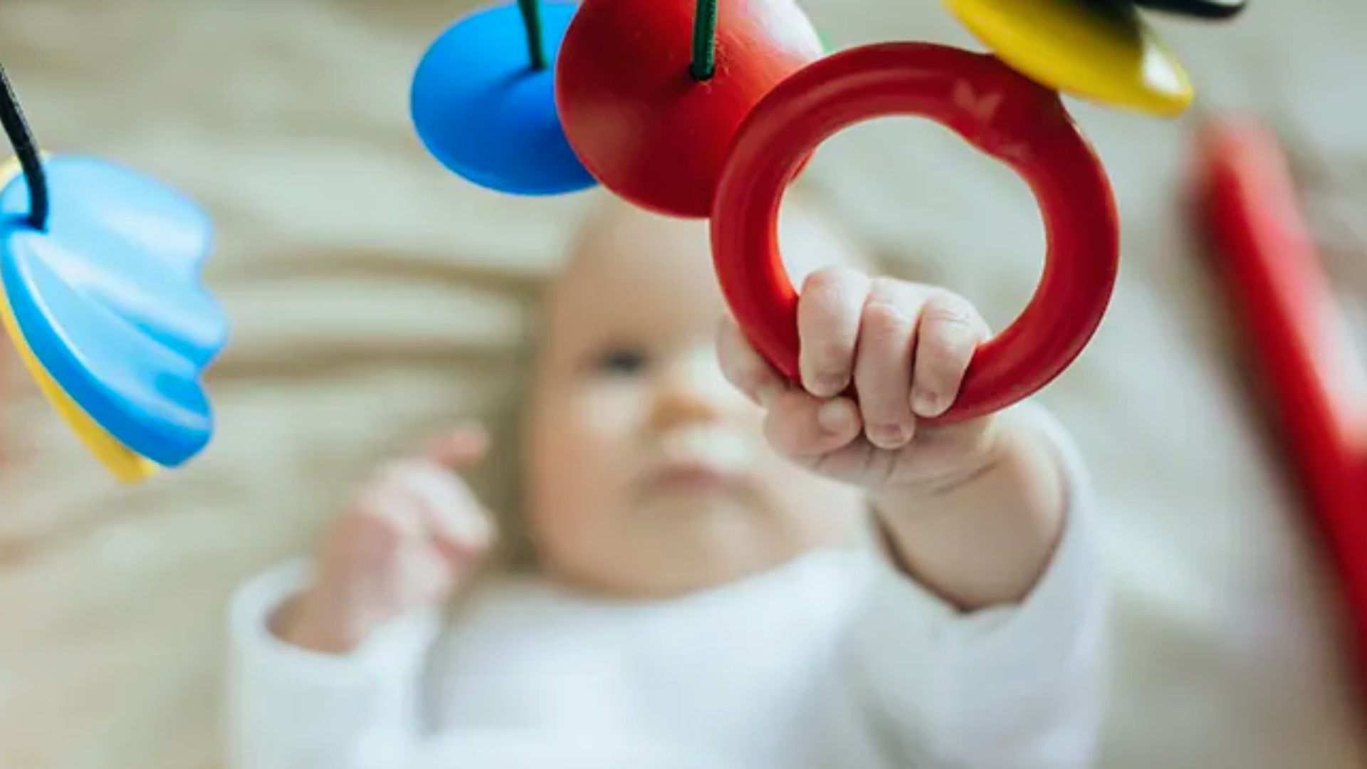 Baby grasping red ring toy from colorful hanging mobile while lying down