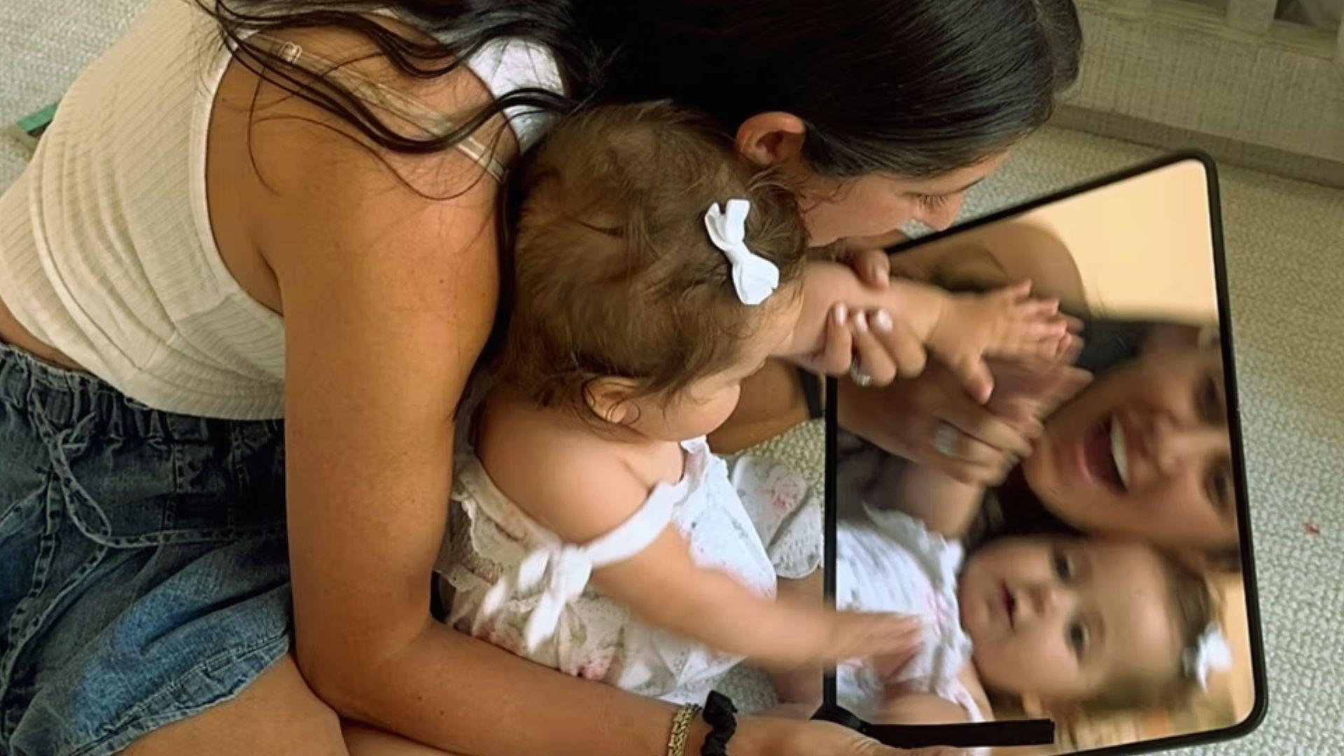 Mother and baby girl with white bow looking at mirror reflection together on nursery floor
