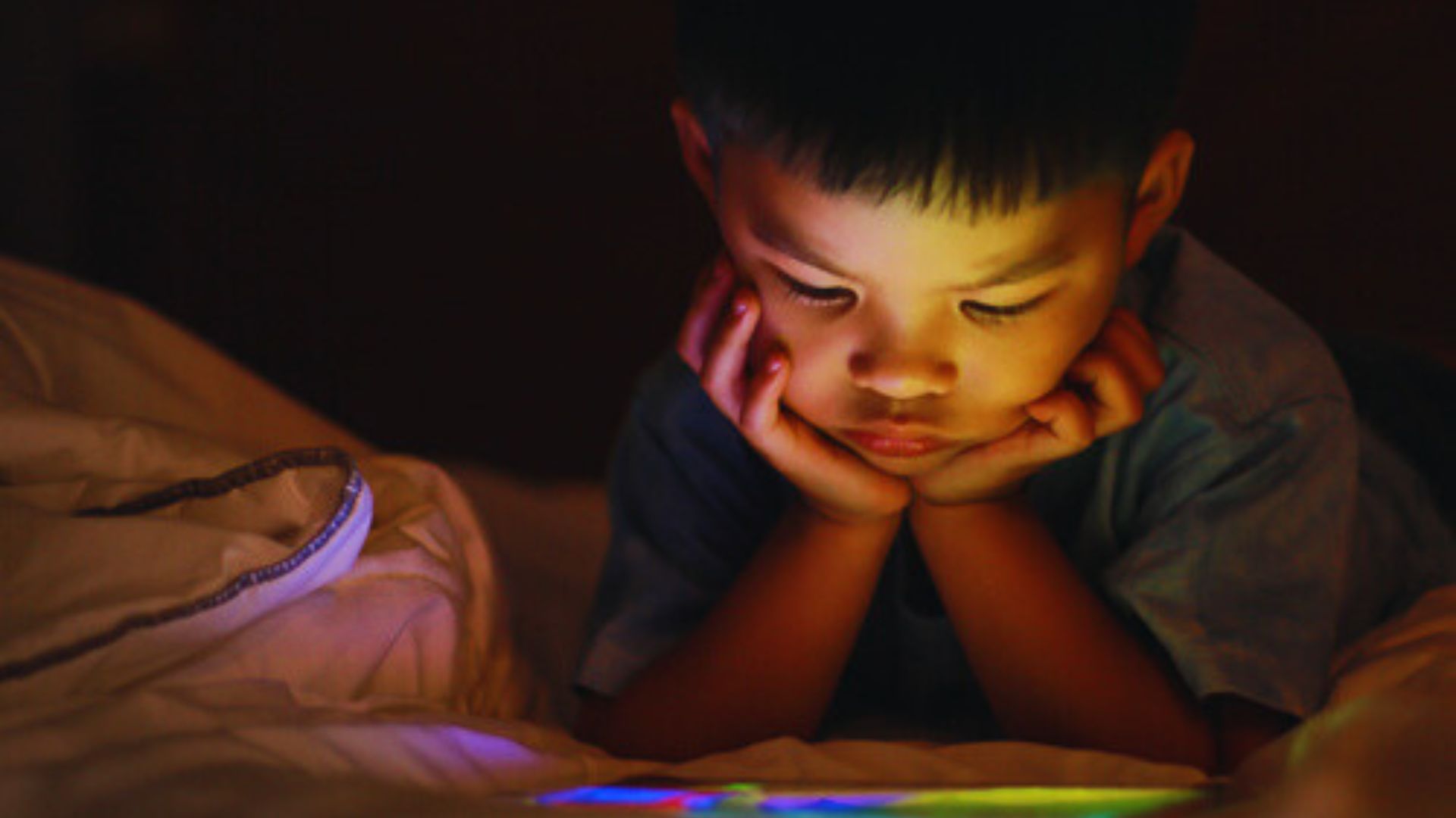 A young boy lying in bed with his hands on his face, reflecting on the importance of limiting screen time before sleep.