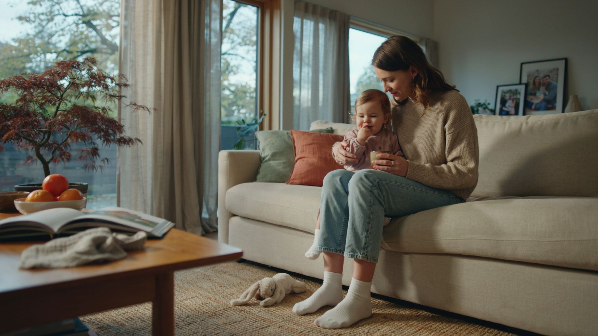 Mother comforting toddler sucking thumb in a cozy living room.