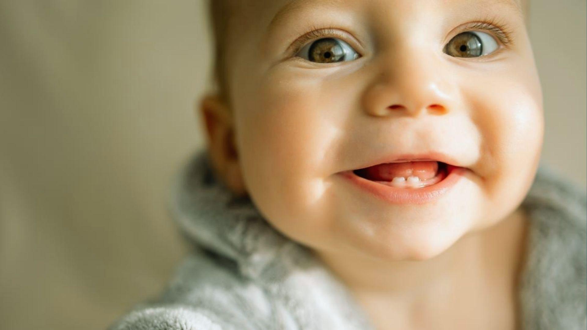Happy baby with brown eyes smiling, showing two tiny lower teeth in gray outfit