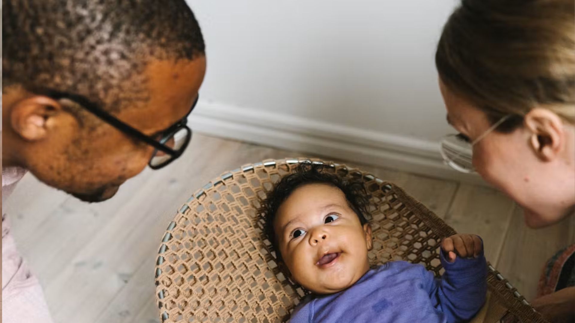 Parents leaning over woven rattan baby bouncer looking at happy baby in blue shirt, close-up bonding moment on wood floor.