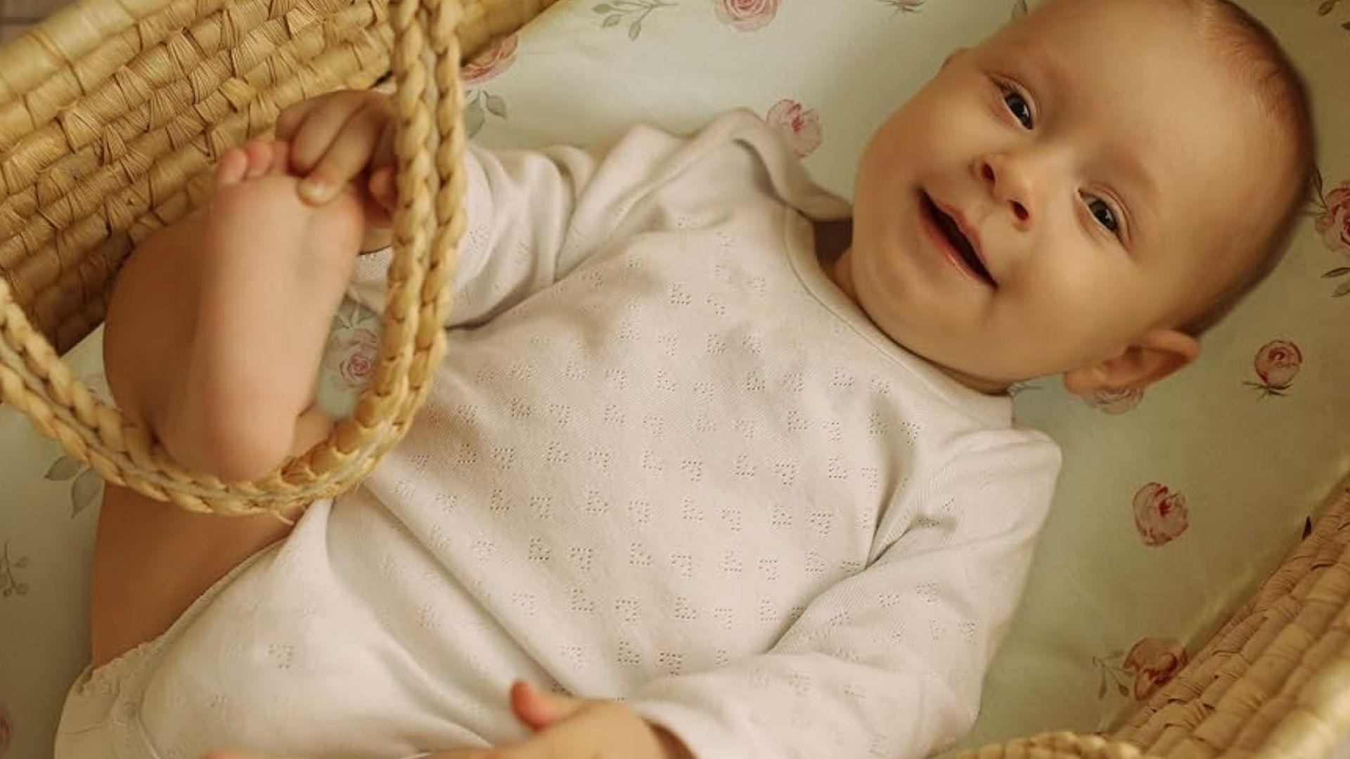 Baby in white onesie lying in woven bassinet on floral sheet holding foot and handle