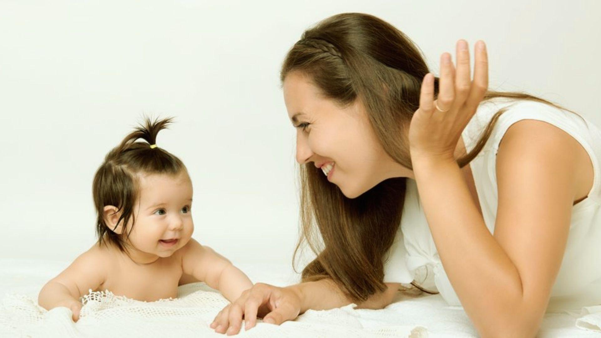 Smiling mother playing with happy baby girl with small ponytail during tummy time on white blanket