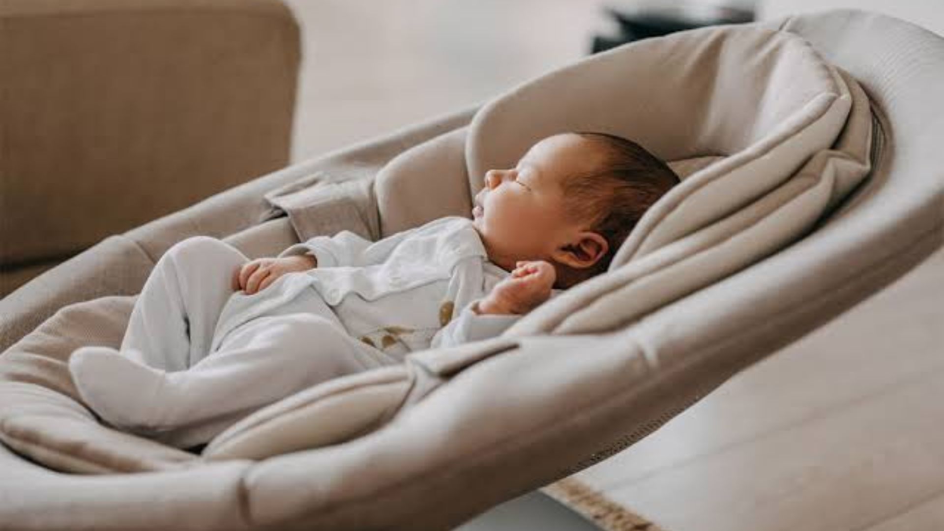 Newborn baby in white outfit sleeping peacefully in plush beige cushioned baby bouncer seat, side view close-up.