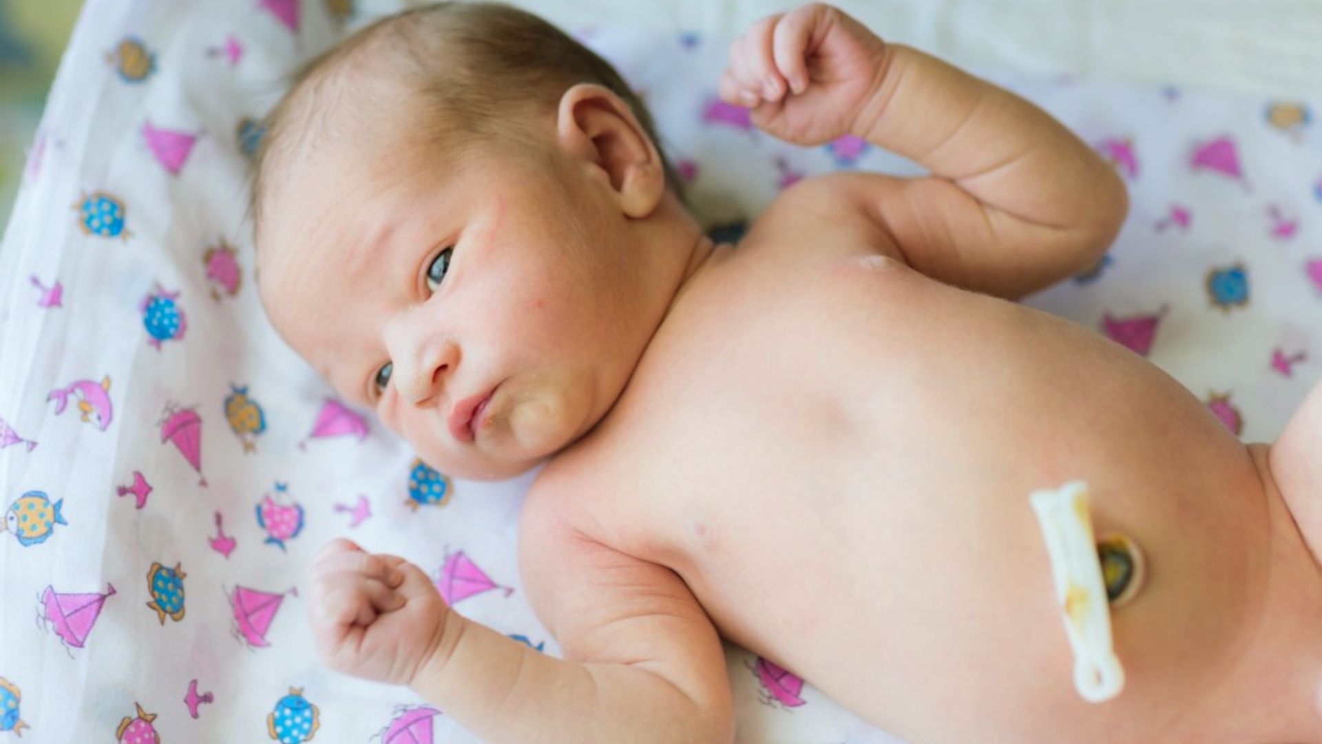 Newborn baby lying on colorful fish-patterned blanket with umbilical cord clamp on belly looking to side