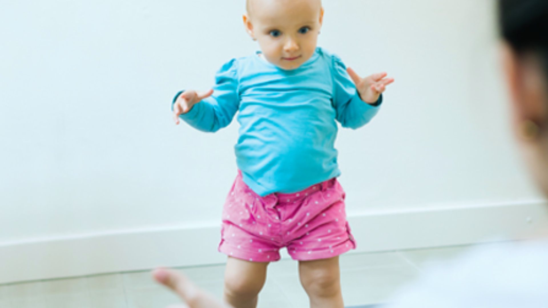 Toddler in blue shirt and pink shorts balancing while learning to walk toward adult indoors